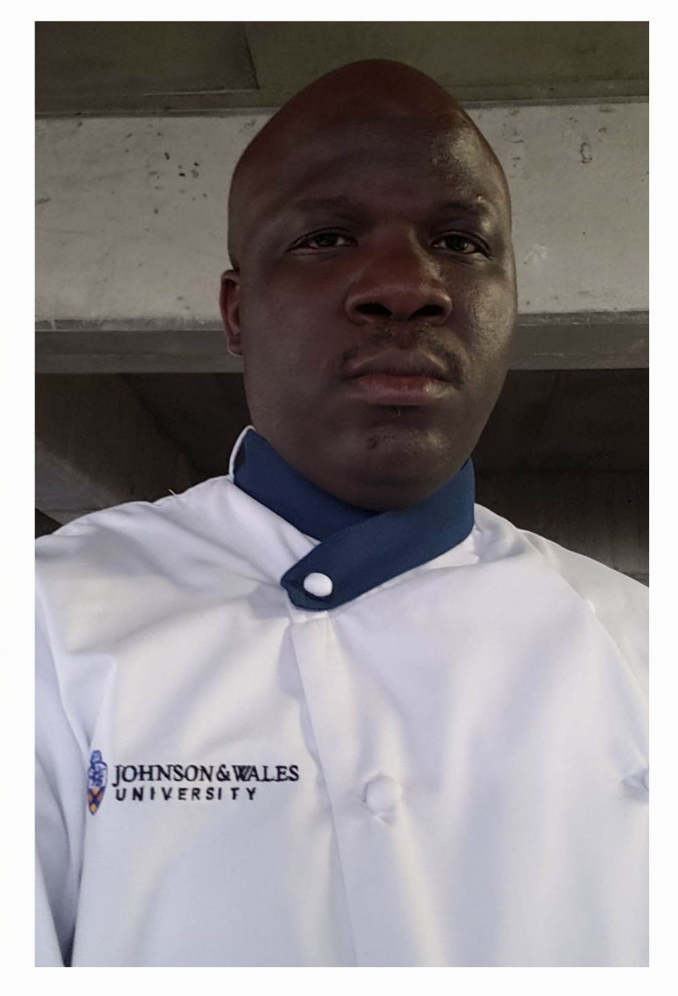 Person wearing a white uniform with a blue collar, standing indoors with a concrete ceiling in the background.