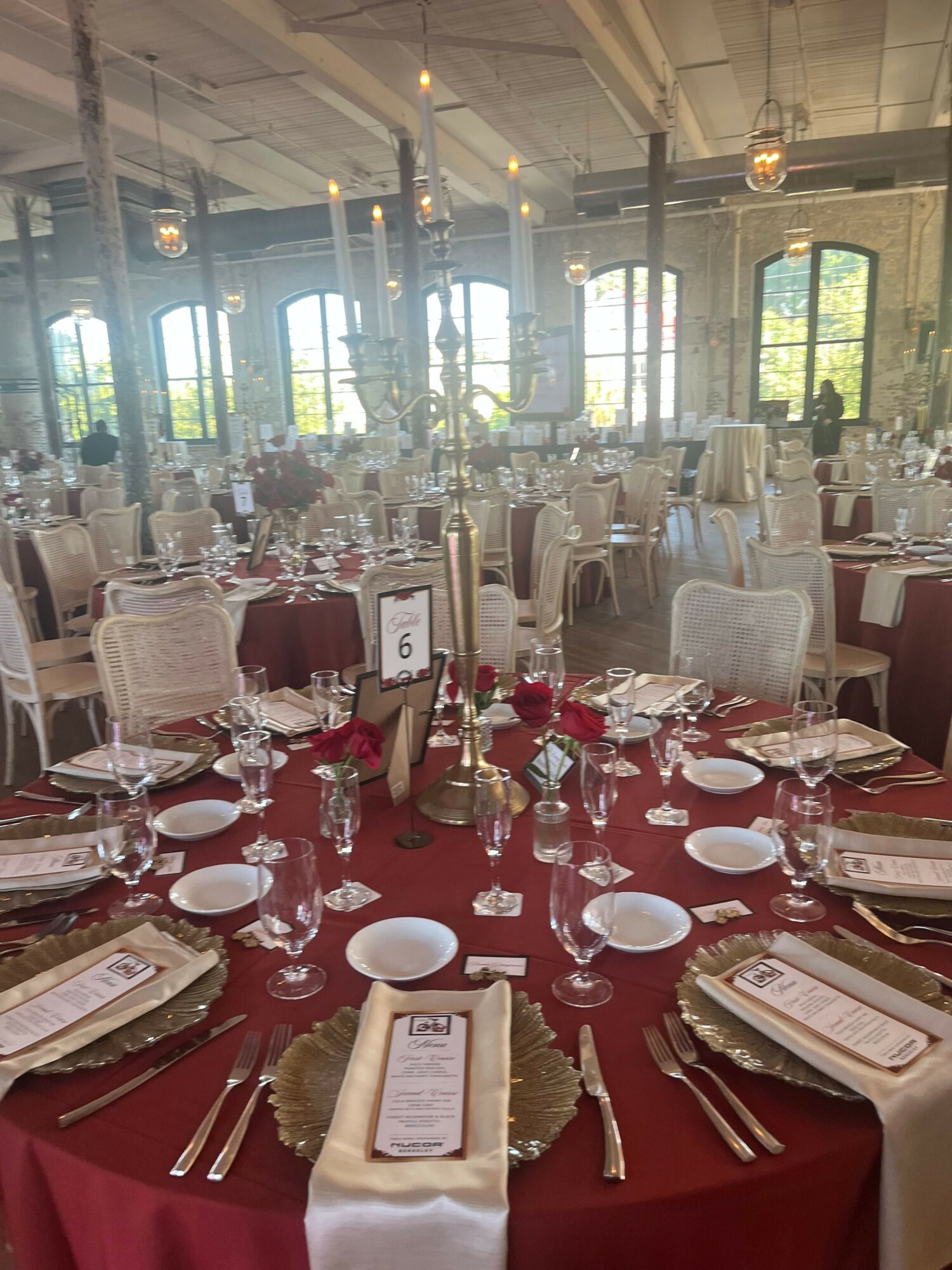 Elegant banquet table set with white plates, glasses, silverware, and floral centerpieces in a bright room with large windows.