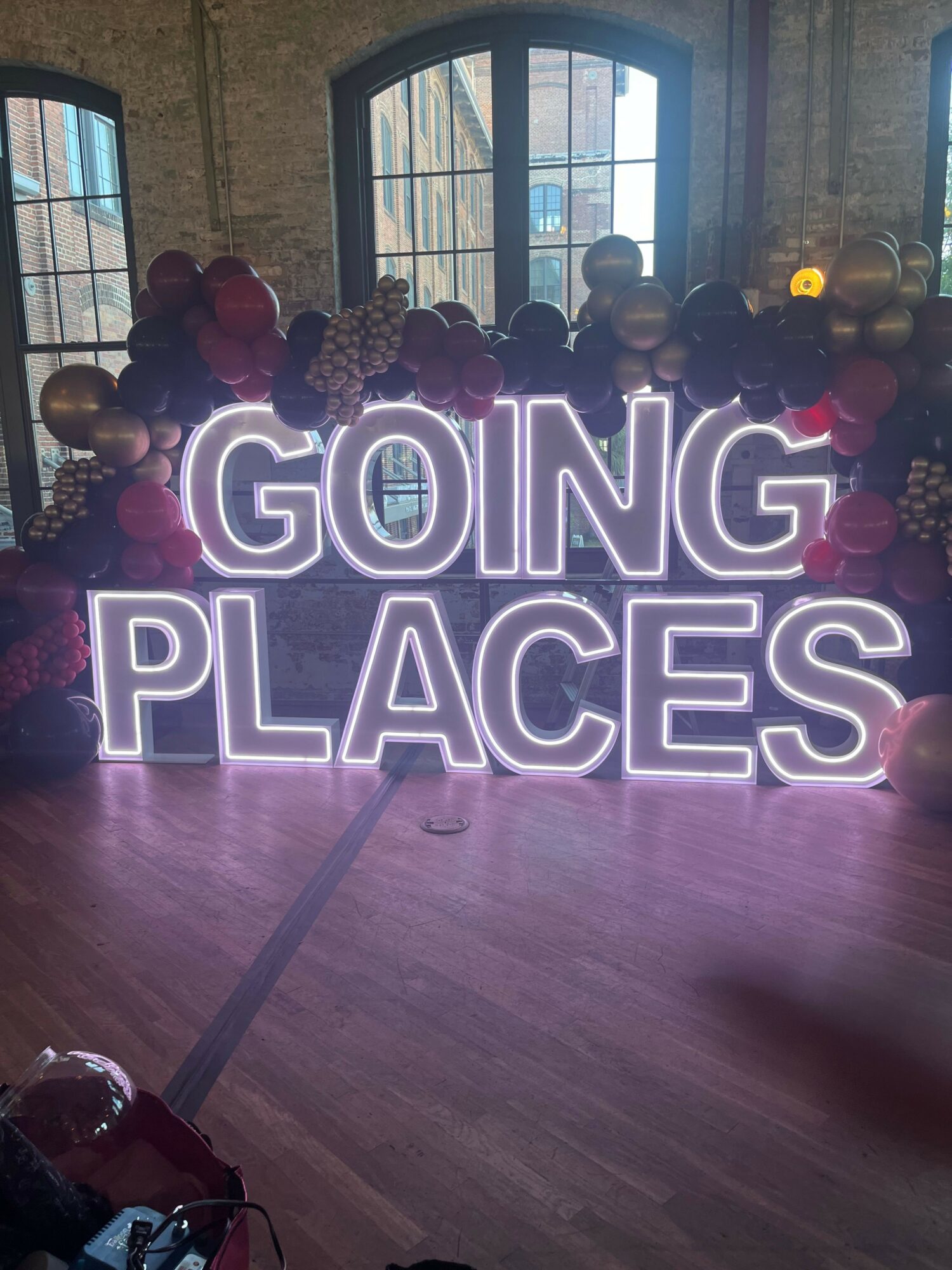 Group of people gathered in a room with large windows, balloons, and a neon sign reading 'GONG PLACES'.