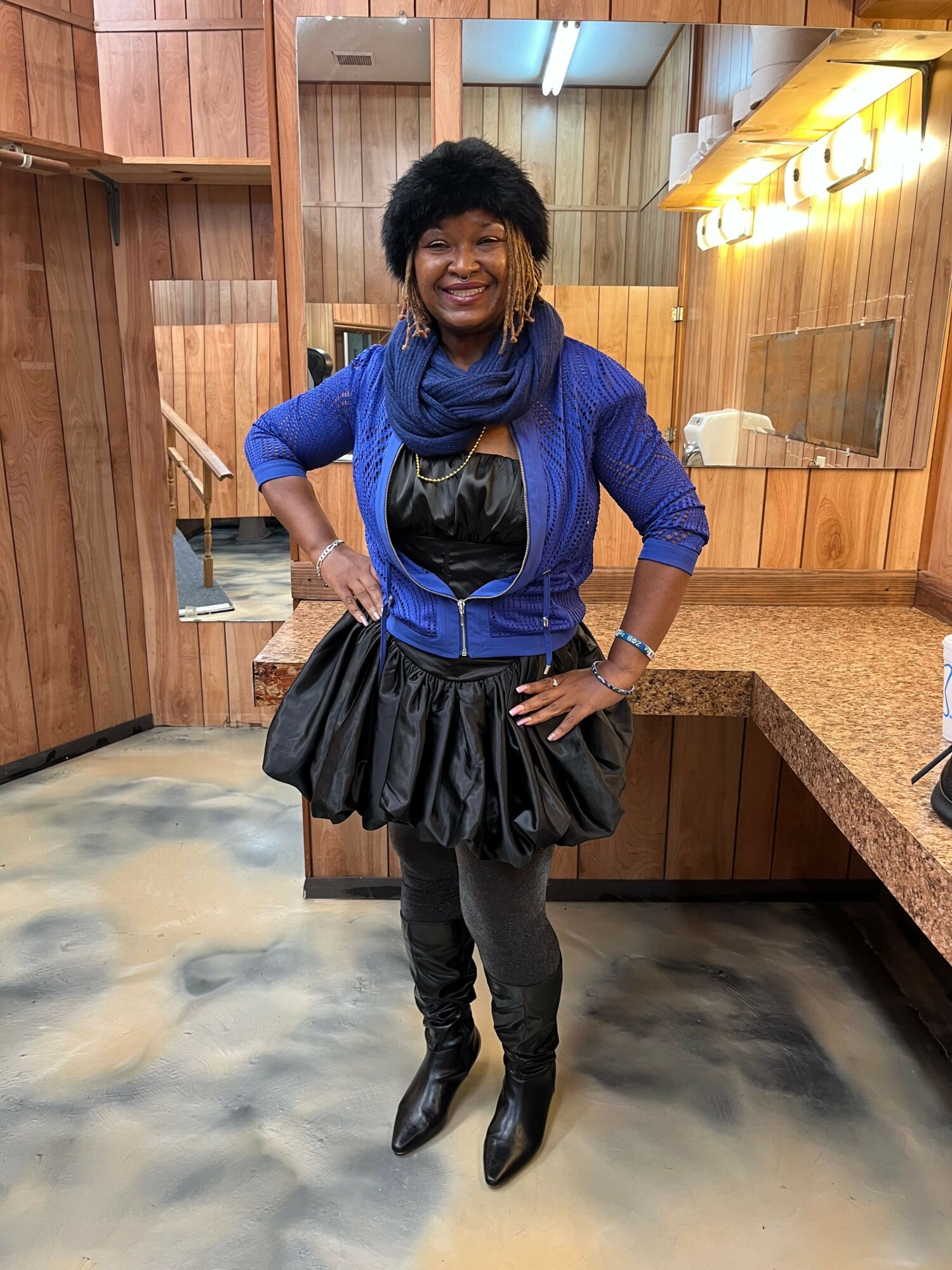 Woman in blue jacket and black skirt standing in a wooden room, holding a phone, with a mirror and lights behind her.