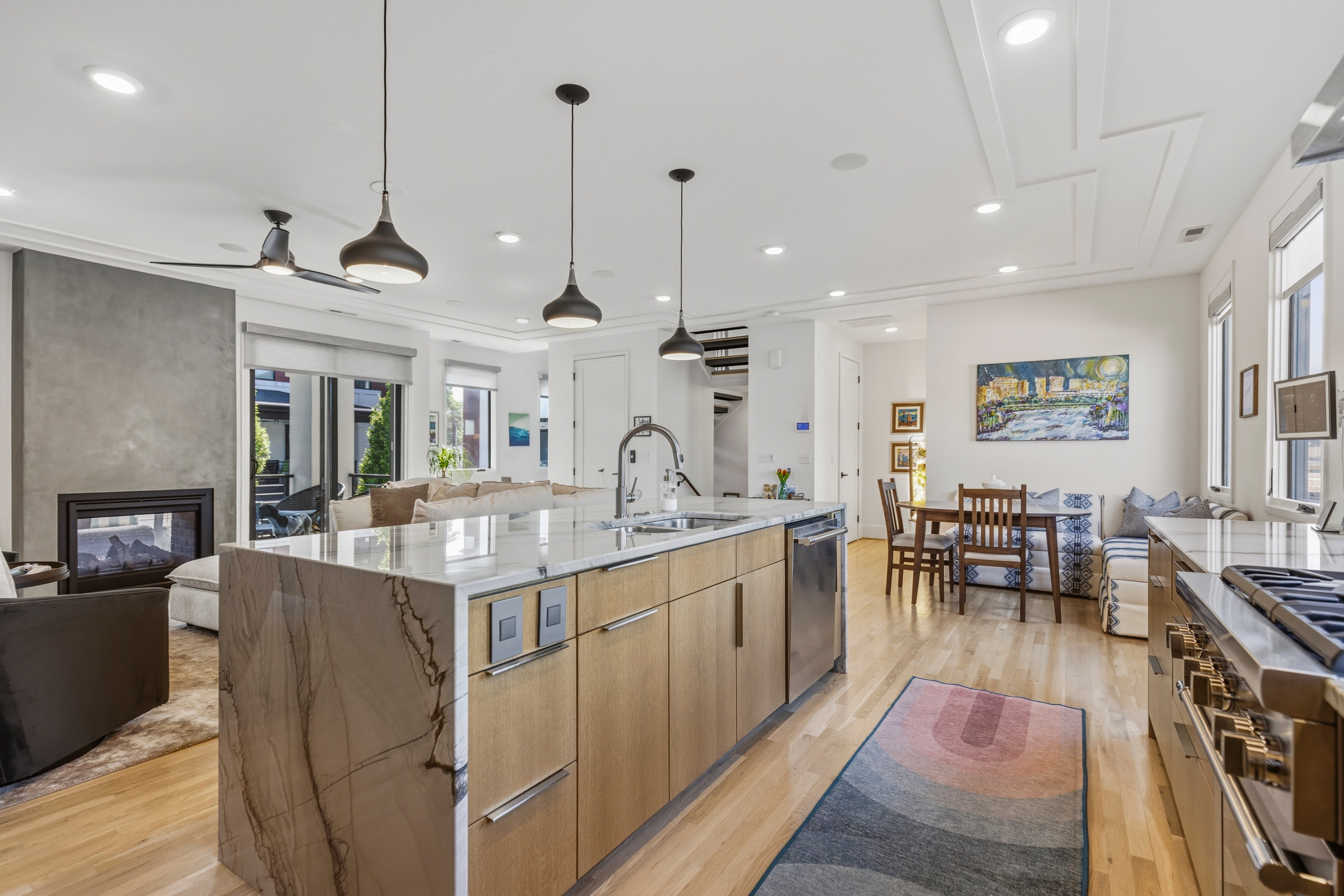 Open-concept kitchen and dining area with island, pendant lights, and large windows, leading to living space with fireplace and artwork.