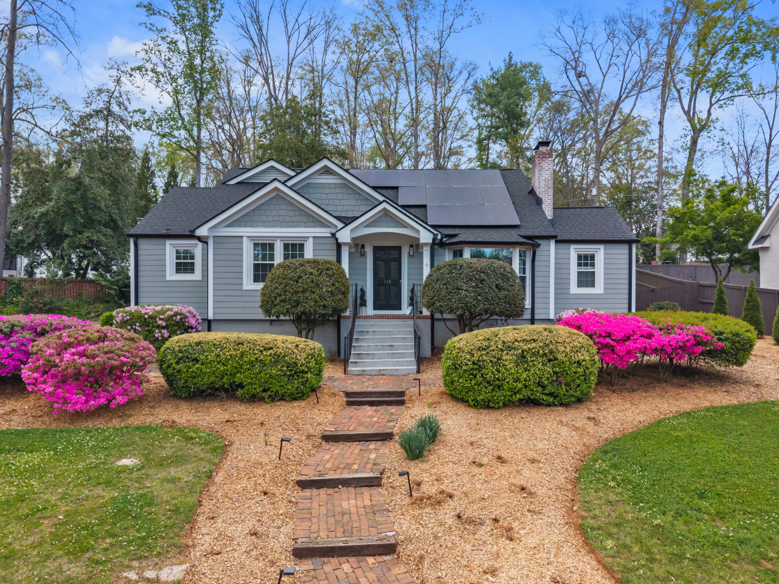 Front view of a house with a pathway, bushes, and flowering plants, surrounded by trees, under a blue sky.
