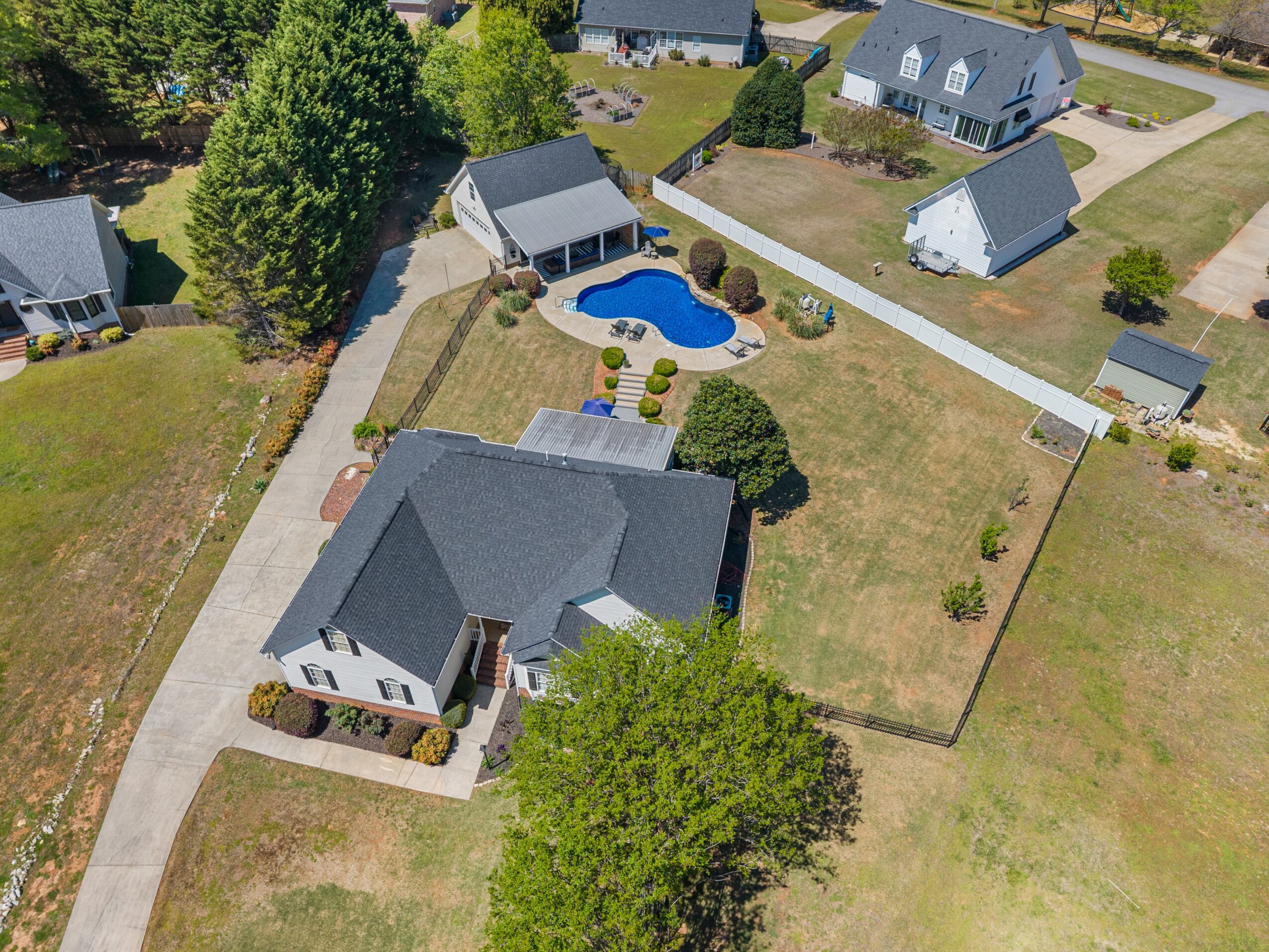 Aerial view of a house with a backyard pool, surrounded by trees and neighboring houses, with a driveway leading to the house.