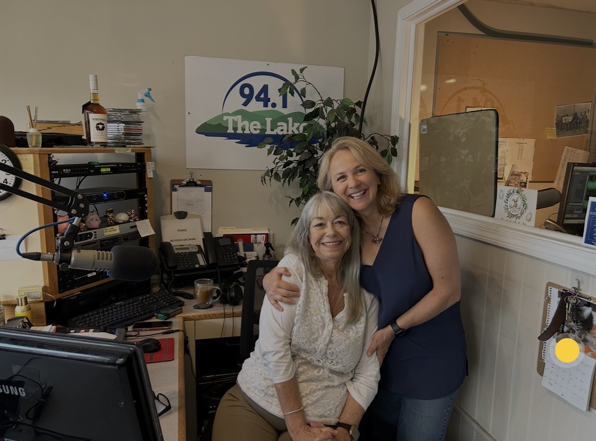Two women smiling and hugging in a radio station studio with equipment and a sign reading 94.1 The Lake.