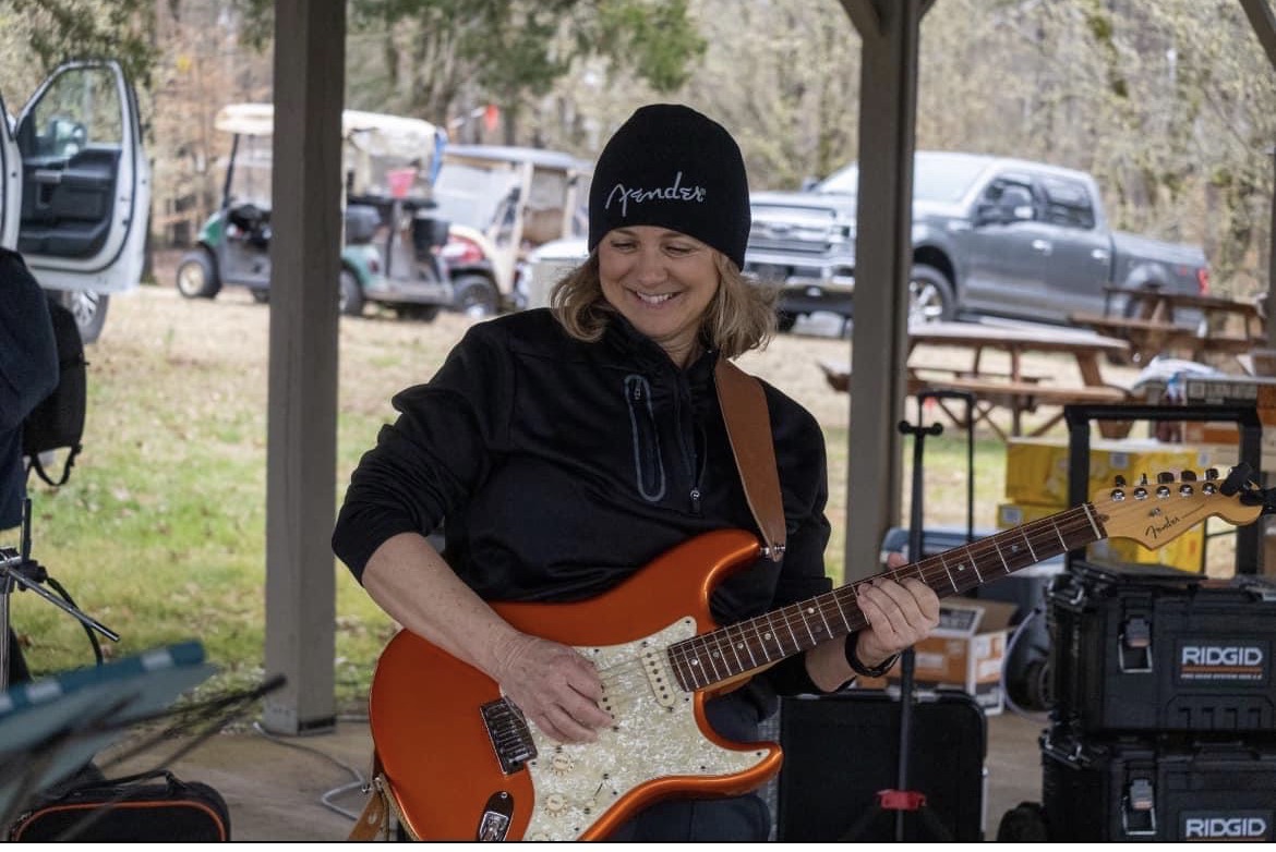 Woman smiling and playing an orange electric guitar outdoors under a shelter, with cars in the background.