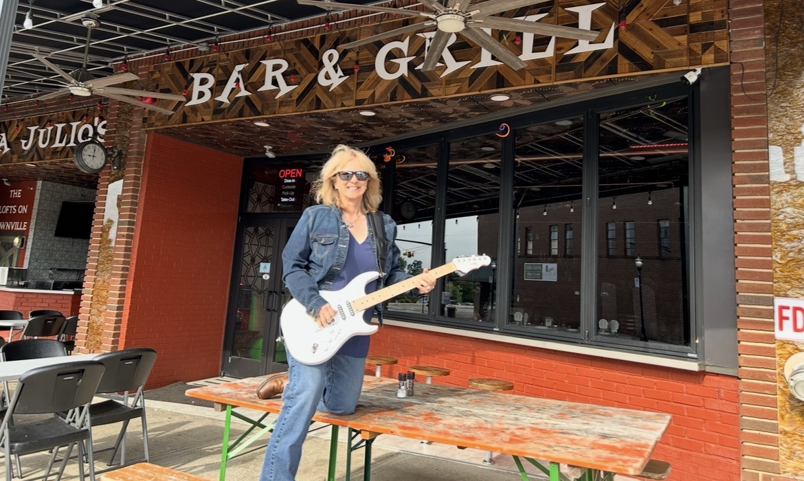 Woman with curly hair and sunglasses holding a white guitar outside a bar and grill, with outdoor seating and a brick building.