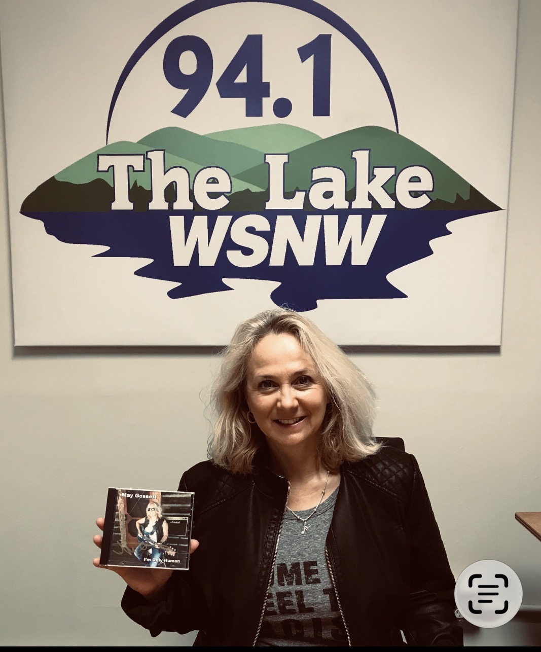 Woman with blonde hair holding a CD, smiling, in front of a sign reading 94.1 The Lake WSNW, with a lake and mountains graphic.