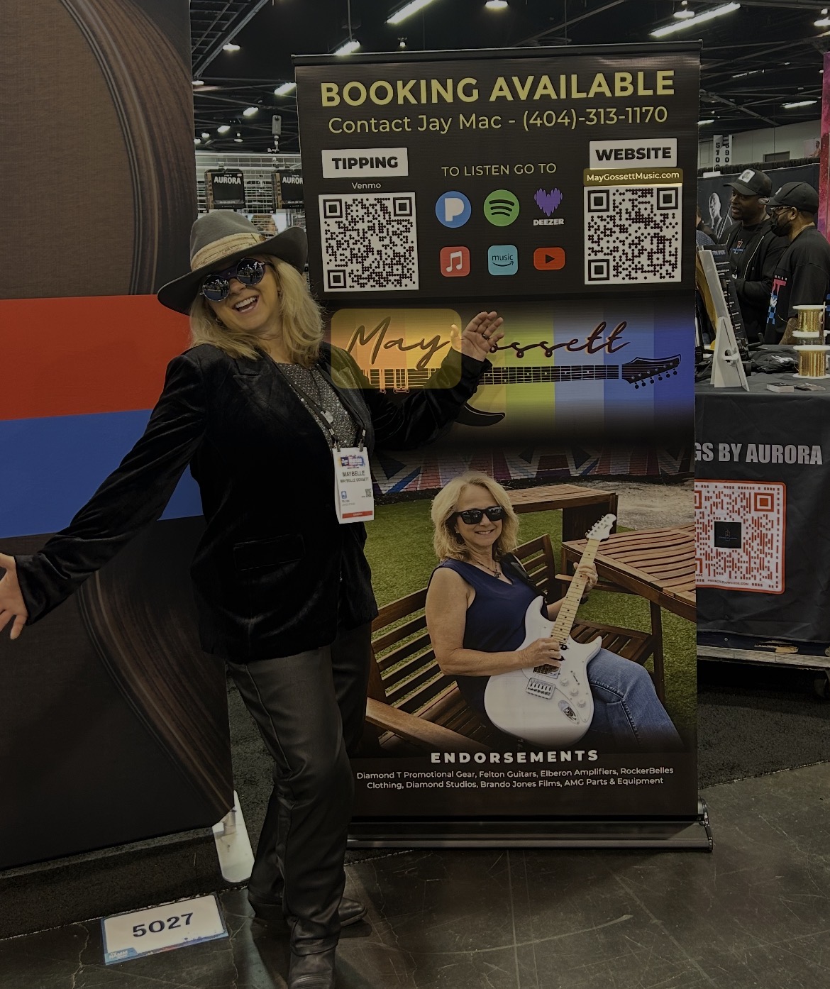 Woman in black outfit and hat standing next to a sign with a photo of herself holding a guitar, at an event.