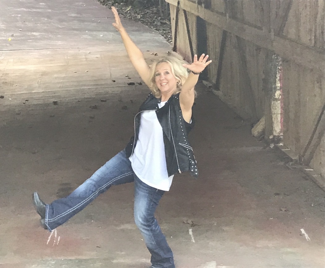 Woman with blonde hair smiling, arms raised, standing on one leg in an outdoor setting with wooden fence behind her.