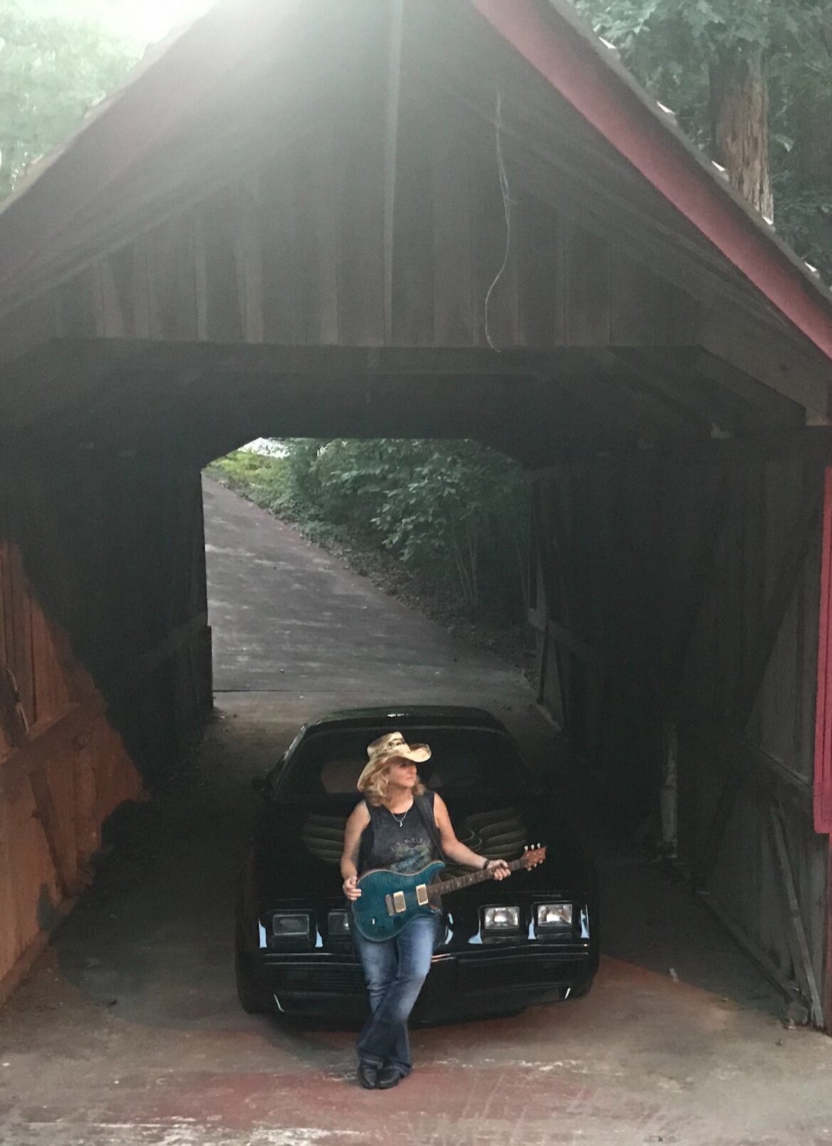Person standing in front of a black car under a wooden structure, holding a guitar, with greenery in the background.