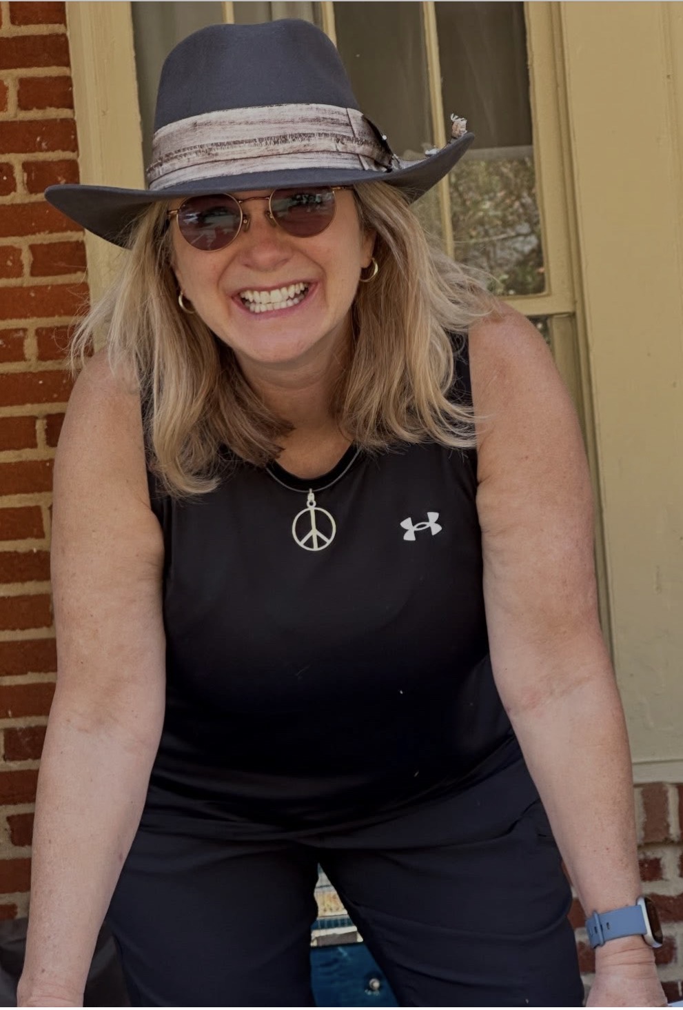 Woman smiling, wearing sunglasses, a hat, and a black sleeveless top with a peace sign necklace, outdoors.