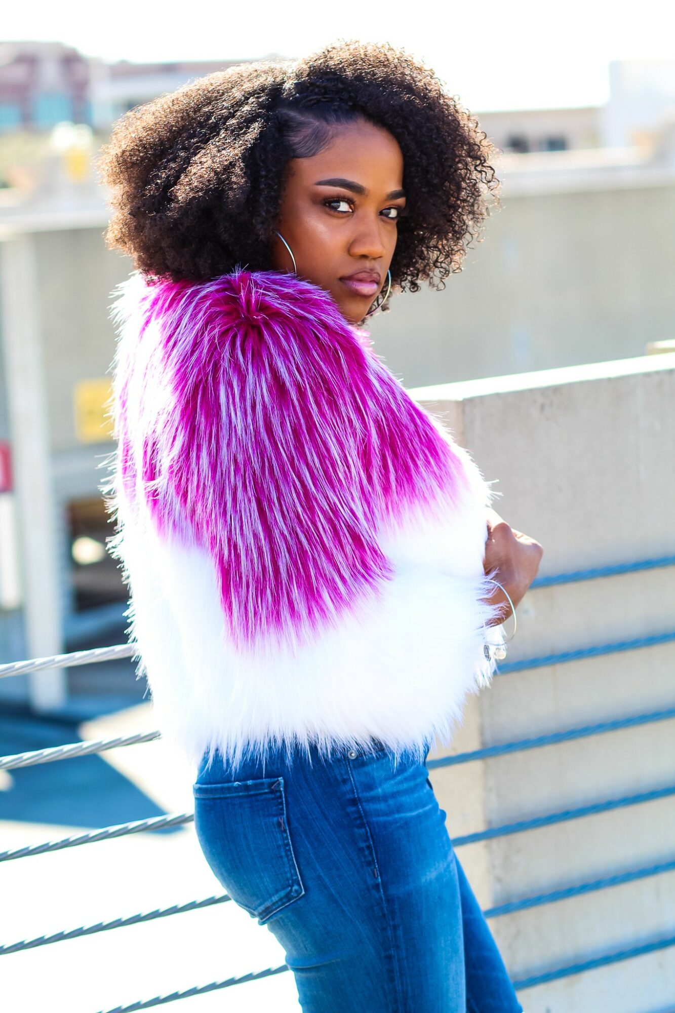 Woman with curly hair wearing a pink and white fur coat, standing outdoors on a balcony railing.