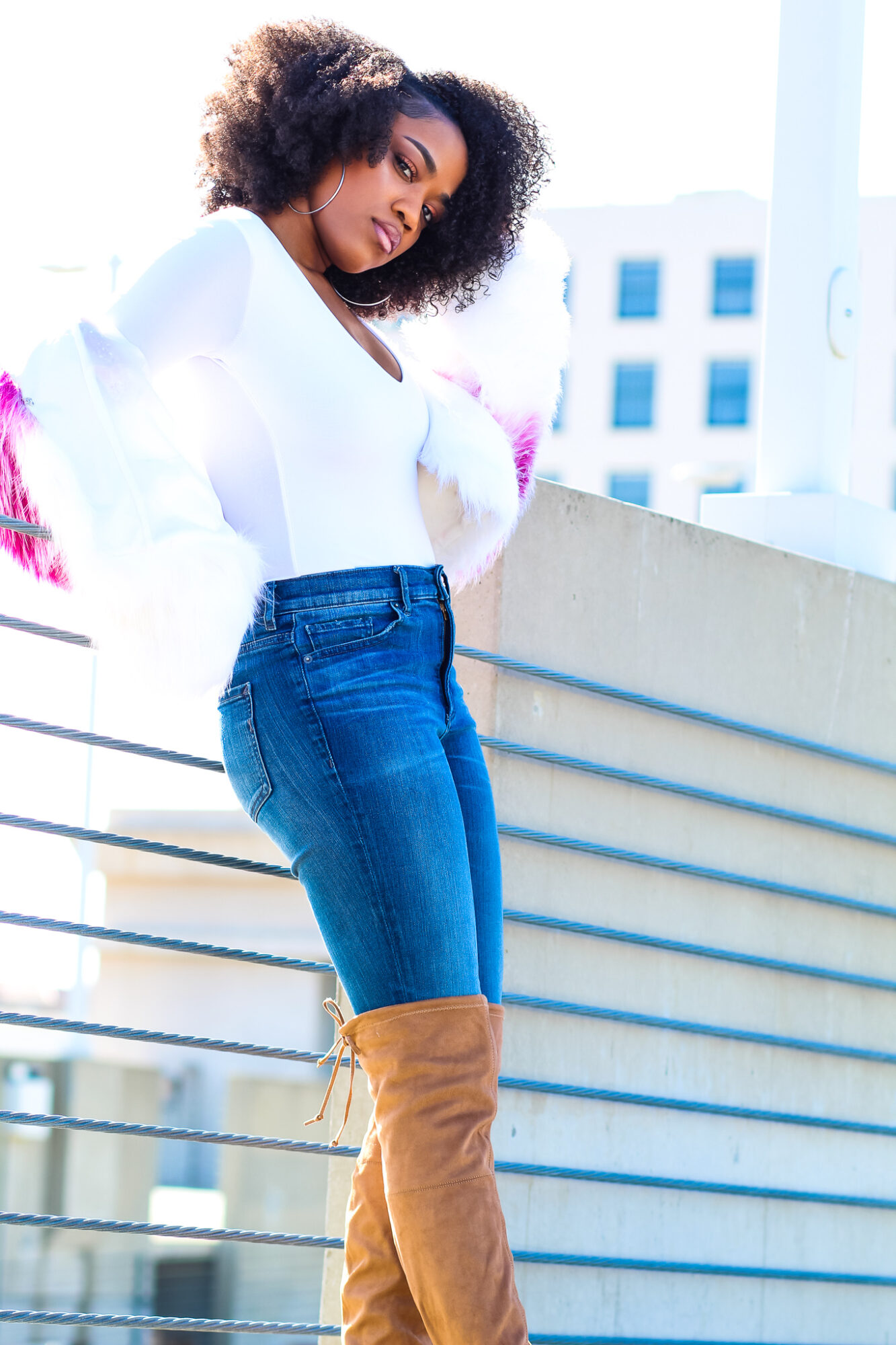 Woman with curly hair leaning on a railing, wearing a white top, blue jeans, and tan boots, with city buildings in background.