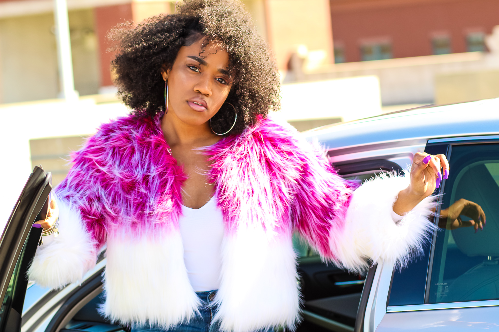 Woman with curly hair wearing a pink and white fur coat, standing next to a car outdoors.