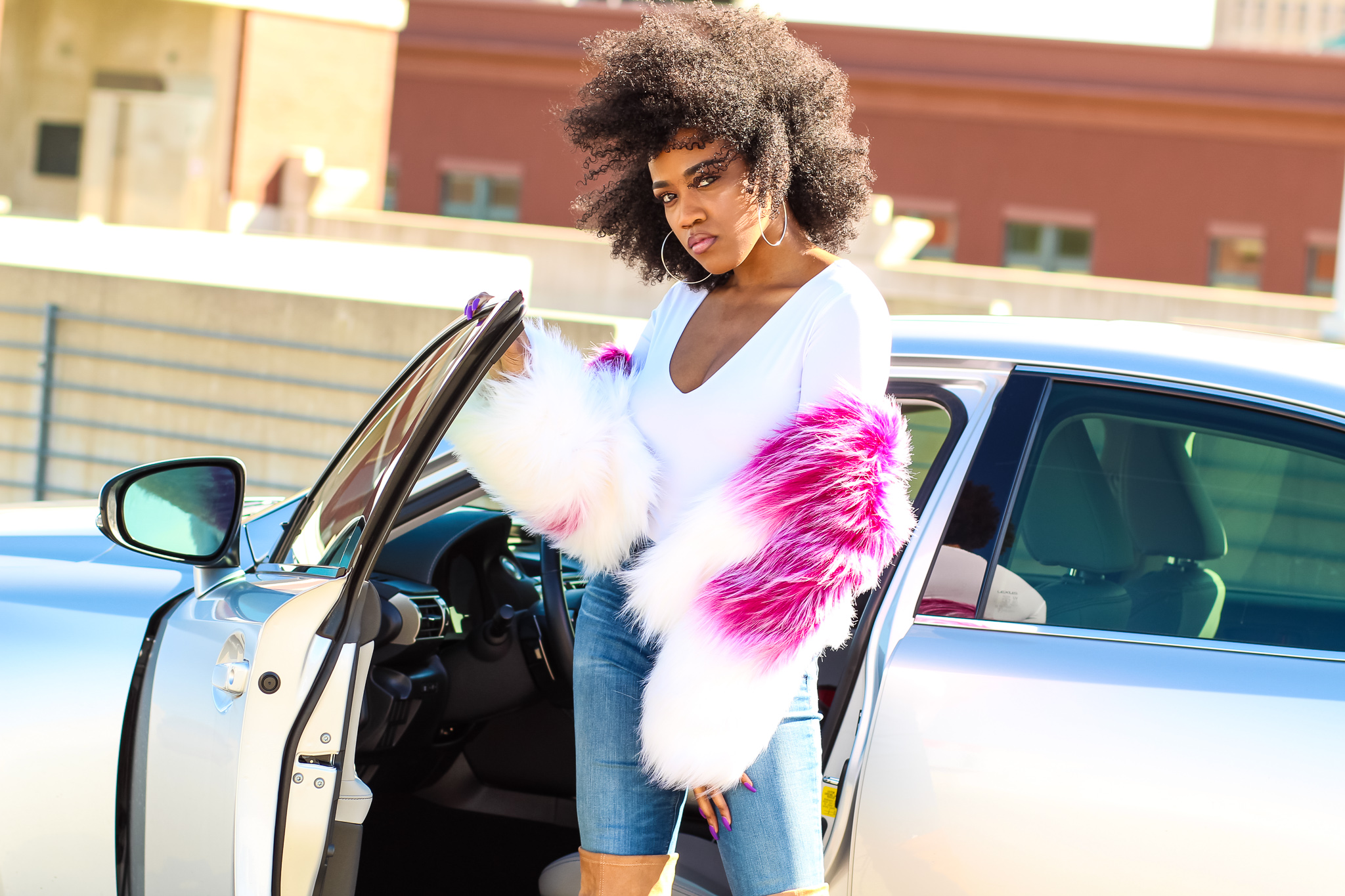 Woman with curly hair wearing a white top and pink fur coat, standing next to a silver car, open door visible.