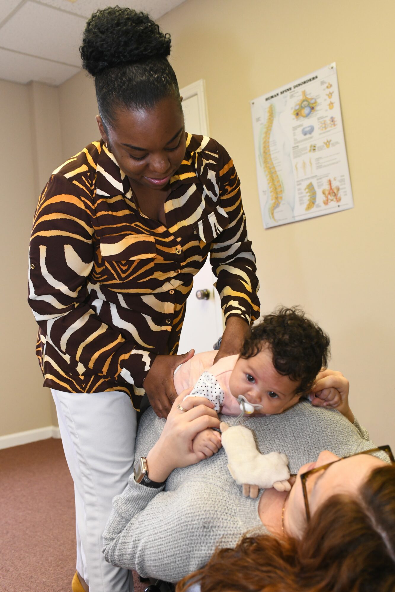 Woman holding a baby with a medical device, smiling, in a room with a poster on the wall.