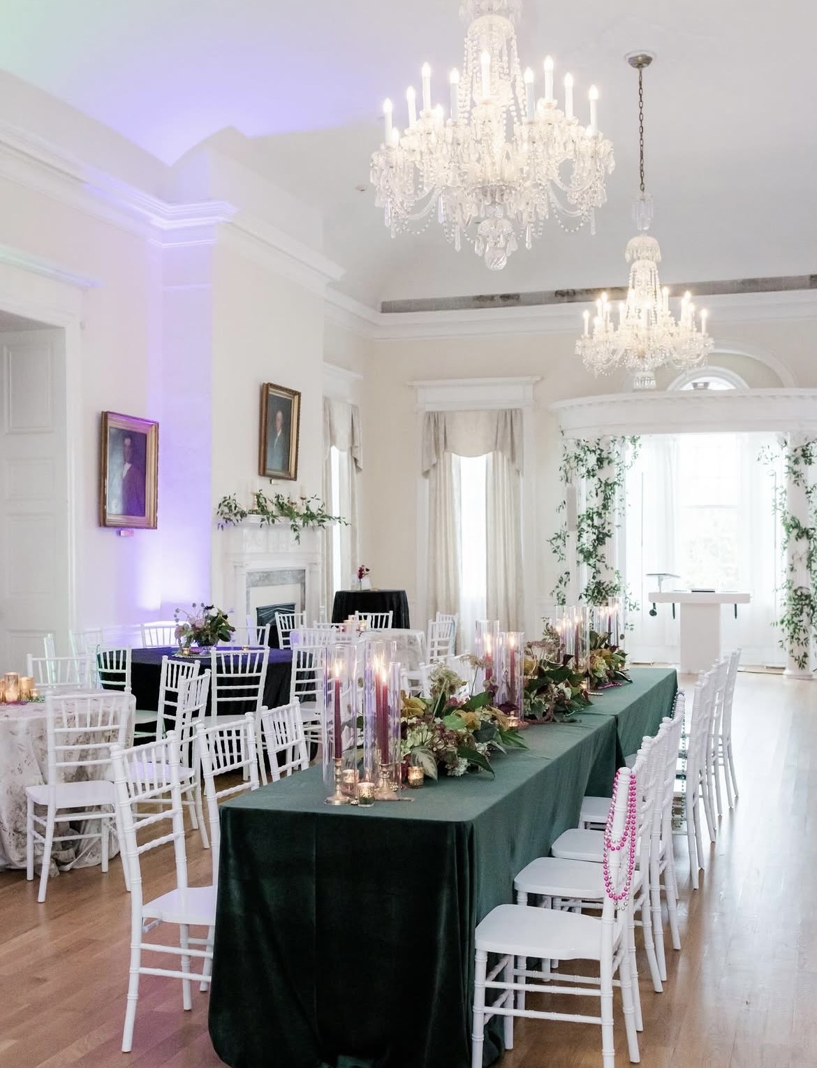Elegant dining room with a long table, white chairs, floral arrangements, chandeliers, and large windows with curtains.