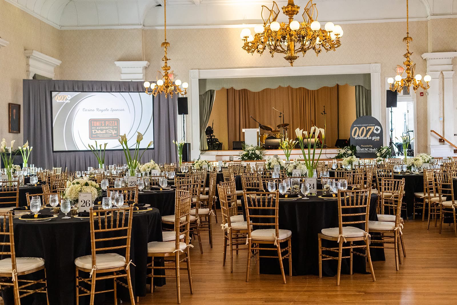 Elegant banquet hall with round tables, black tablecloths, floral centerpieces, and a stage with a large screen and curtains.