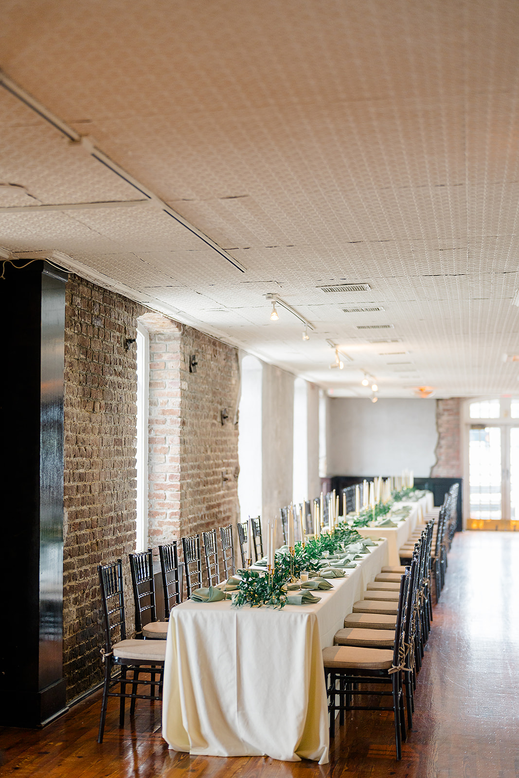 Long dining table with white tablecloth and greenery centerpiece, chairs along both sides, in a room with brick walls and large windows.