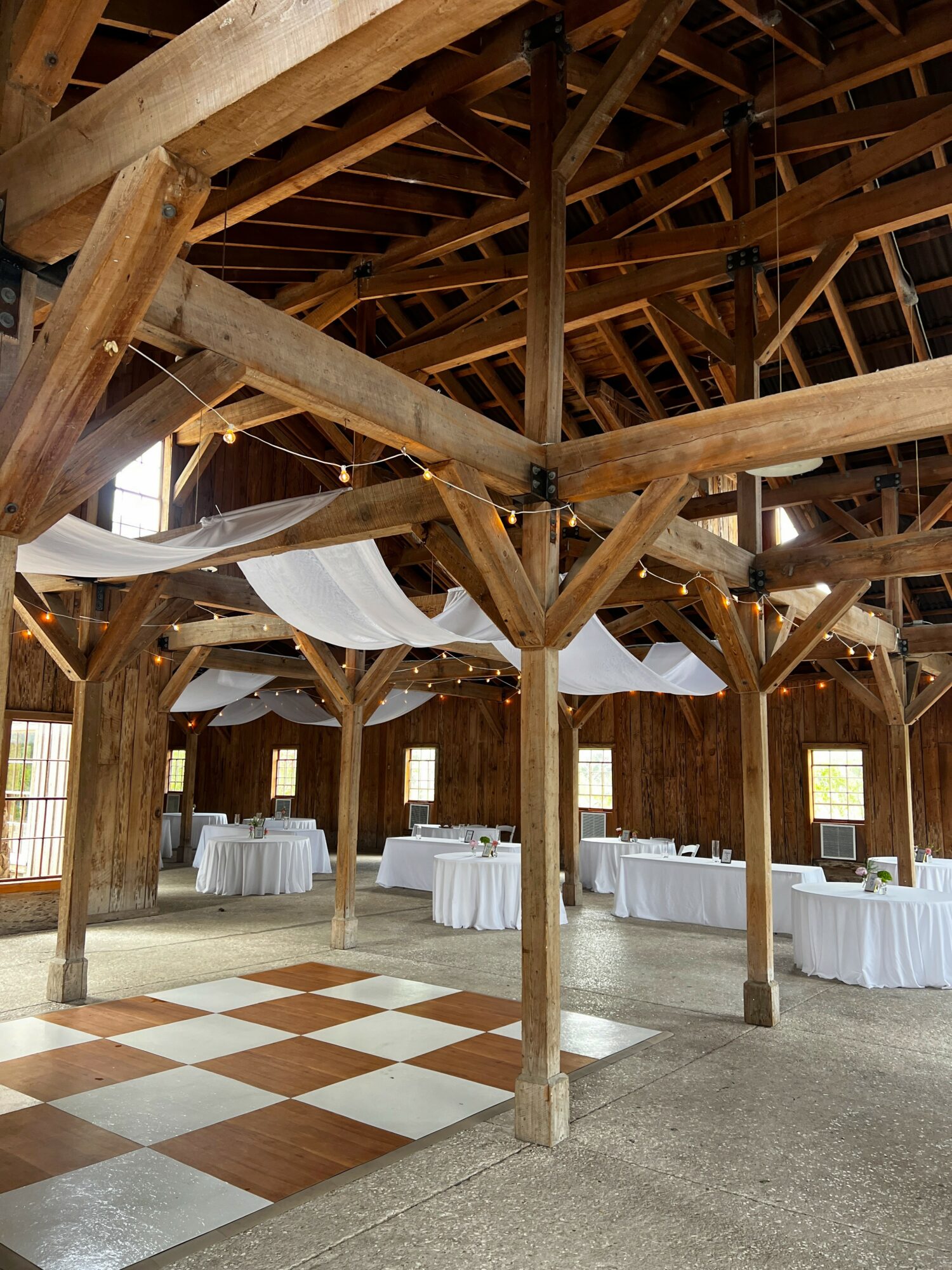 Interior of a wooden barn with tables and white tablecloths, wooden beams, and a checkered dance floor.