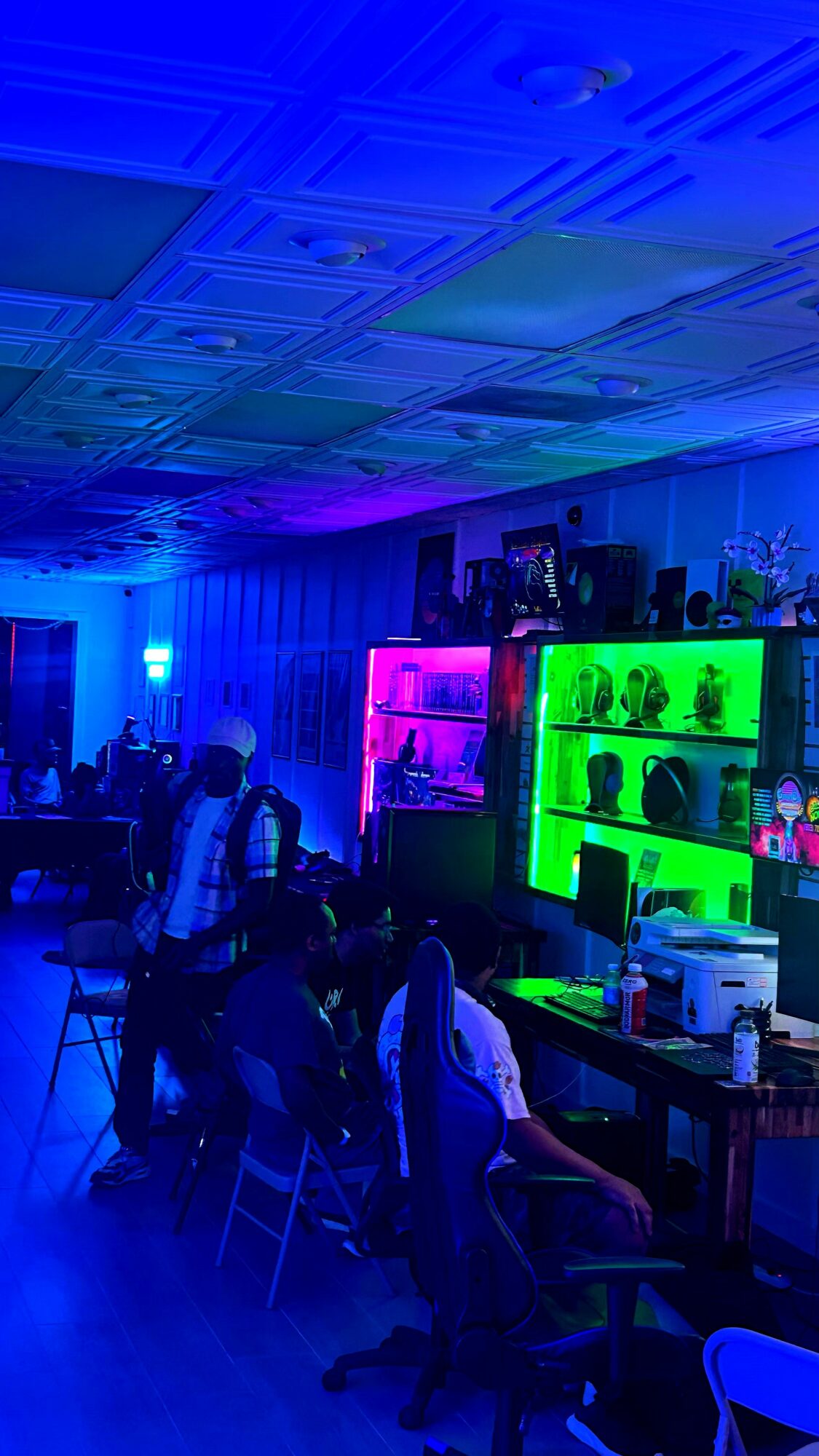 People sitting at tables in a room with colorful illuminated shelves and blue lighting, ceiling with patterned panels.