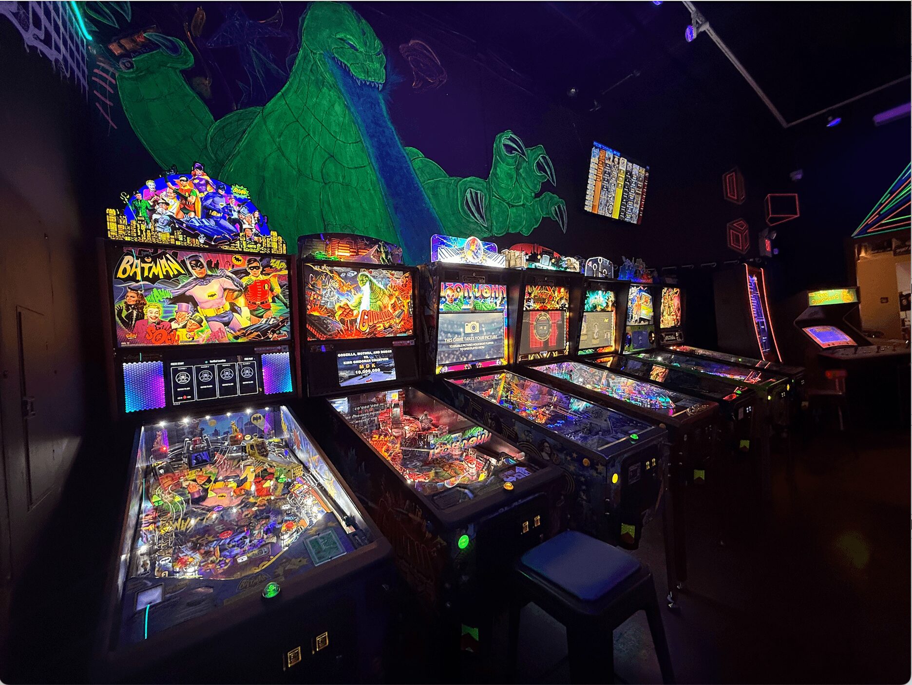 Row of five pinball machines with colorful lights in a dark arcade, with a green dragon mural on the wall behind.