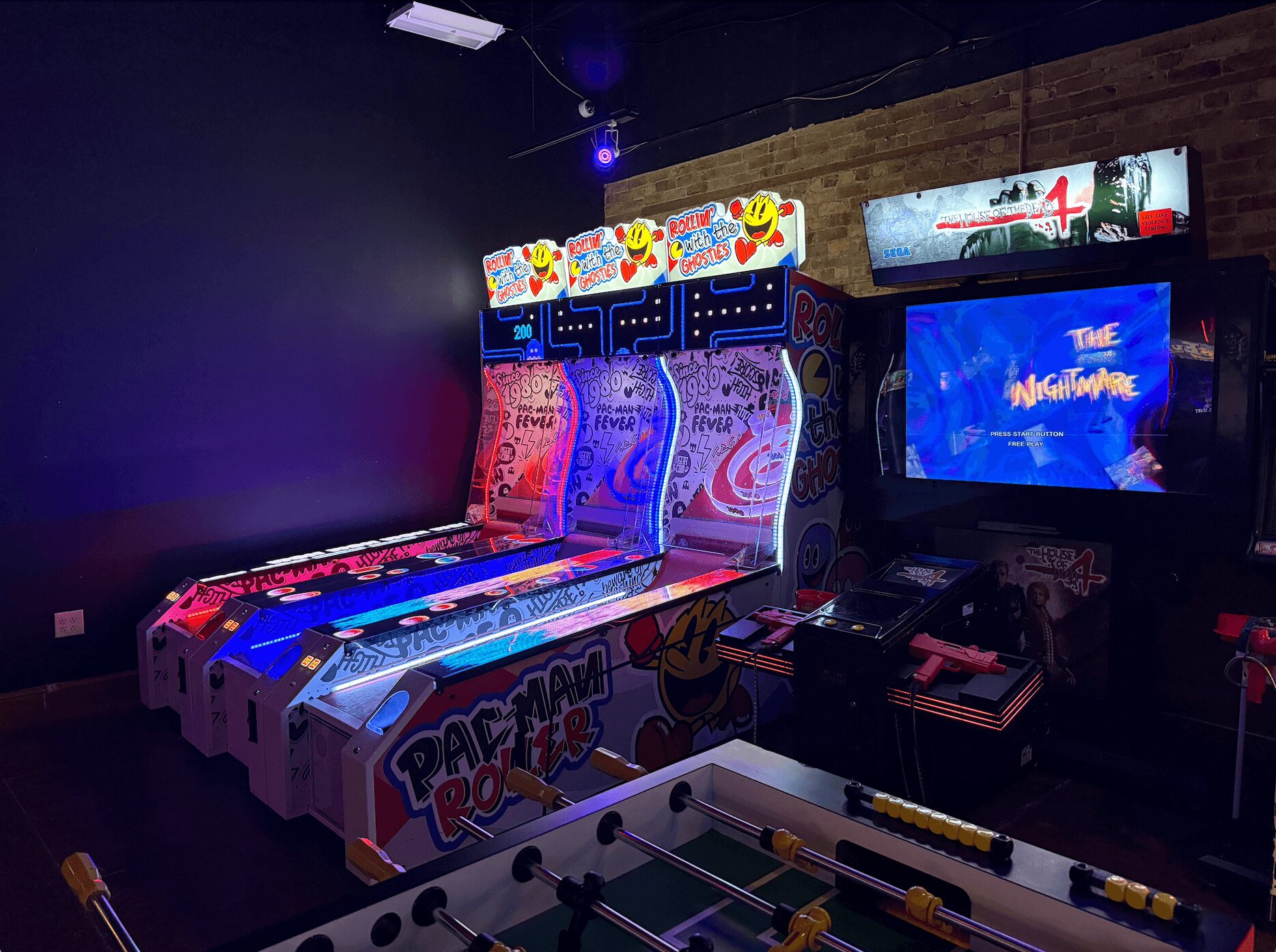 Arcade room with Pac-Man and Donkey Kong machines, and a foosball table in front, dimly lit with colorful game screens.