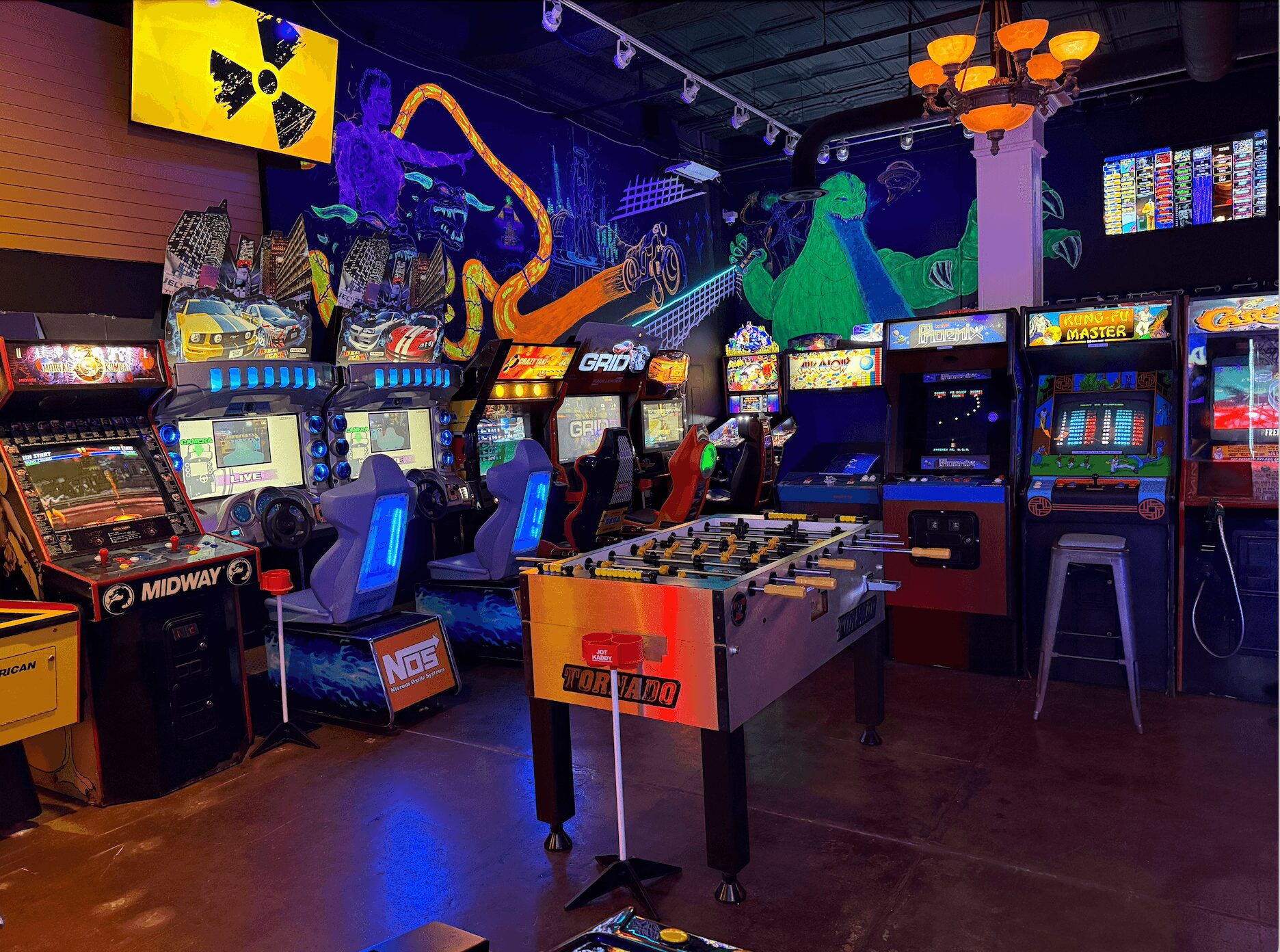 Arcade with colorful game machines, neon lights, and a foosball table in the center, with a mural of dinosaurs on the wall.