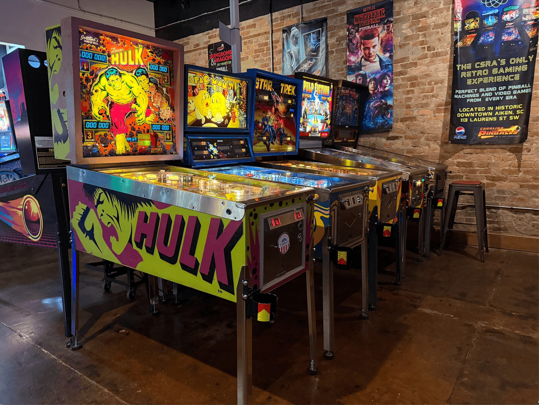 Arcade with pinball machines and video game screens, brick wall background, promotional poster on the wall, wooden floor.