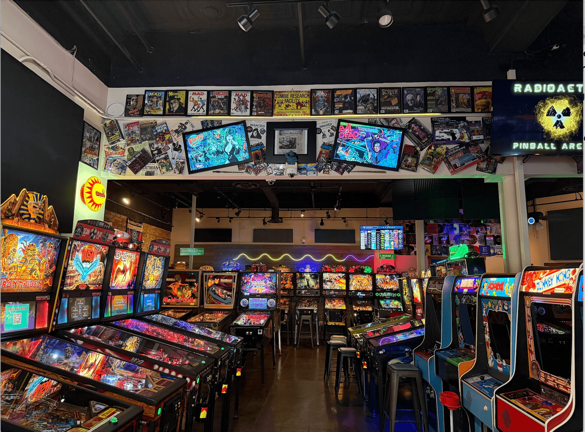 Arcade filled with pinball machines and video games, colorful screens, and a dark ceiling with overhead lights.
