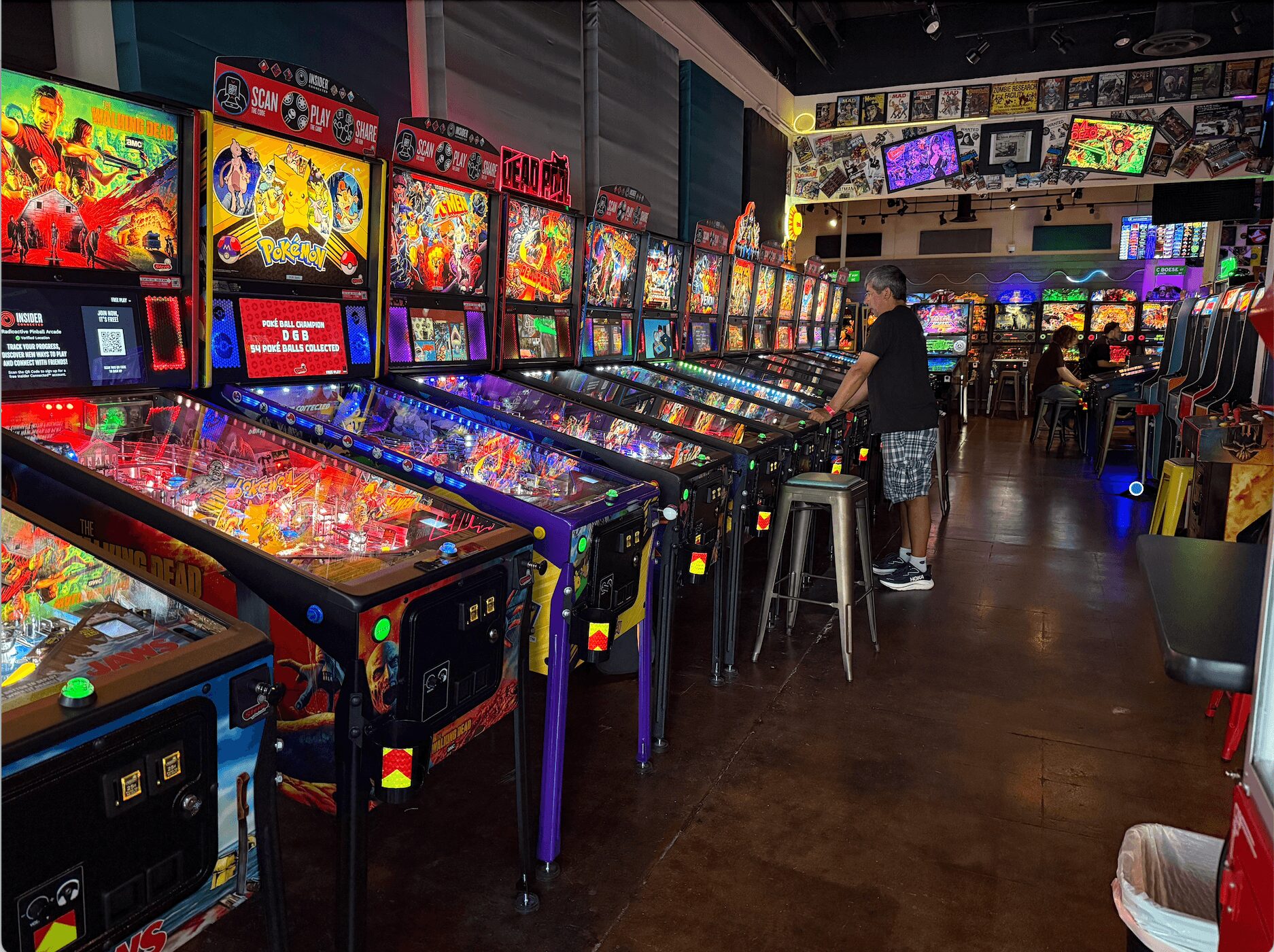 Arcade with multiple pinball machines and a person playing, colorful screens and dim lighting, wooden floor, seating area.
