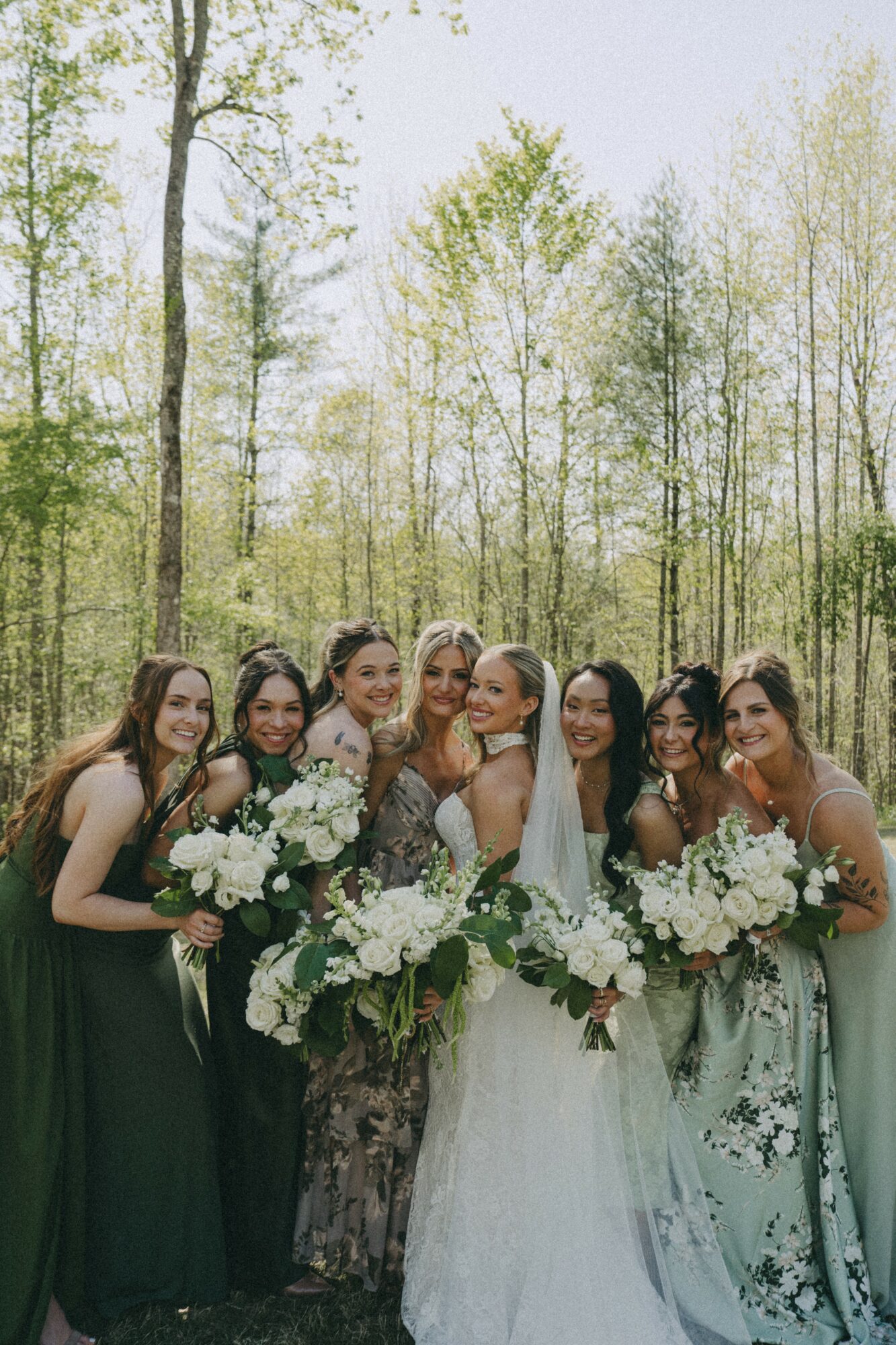 Group of women in wedding dresses and formal attire holding bouquets outdoors in a wooded area.