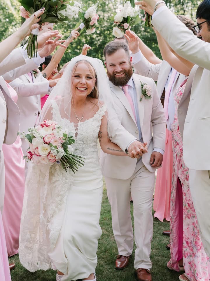 Bride and groom smiling, holding hands, surrounded by friends outdoors, celebrating wedding, with flowers and greenery in background.