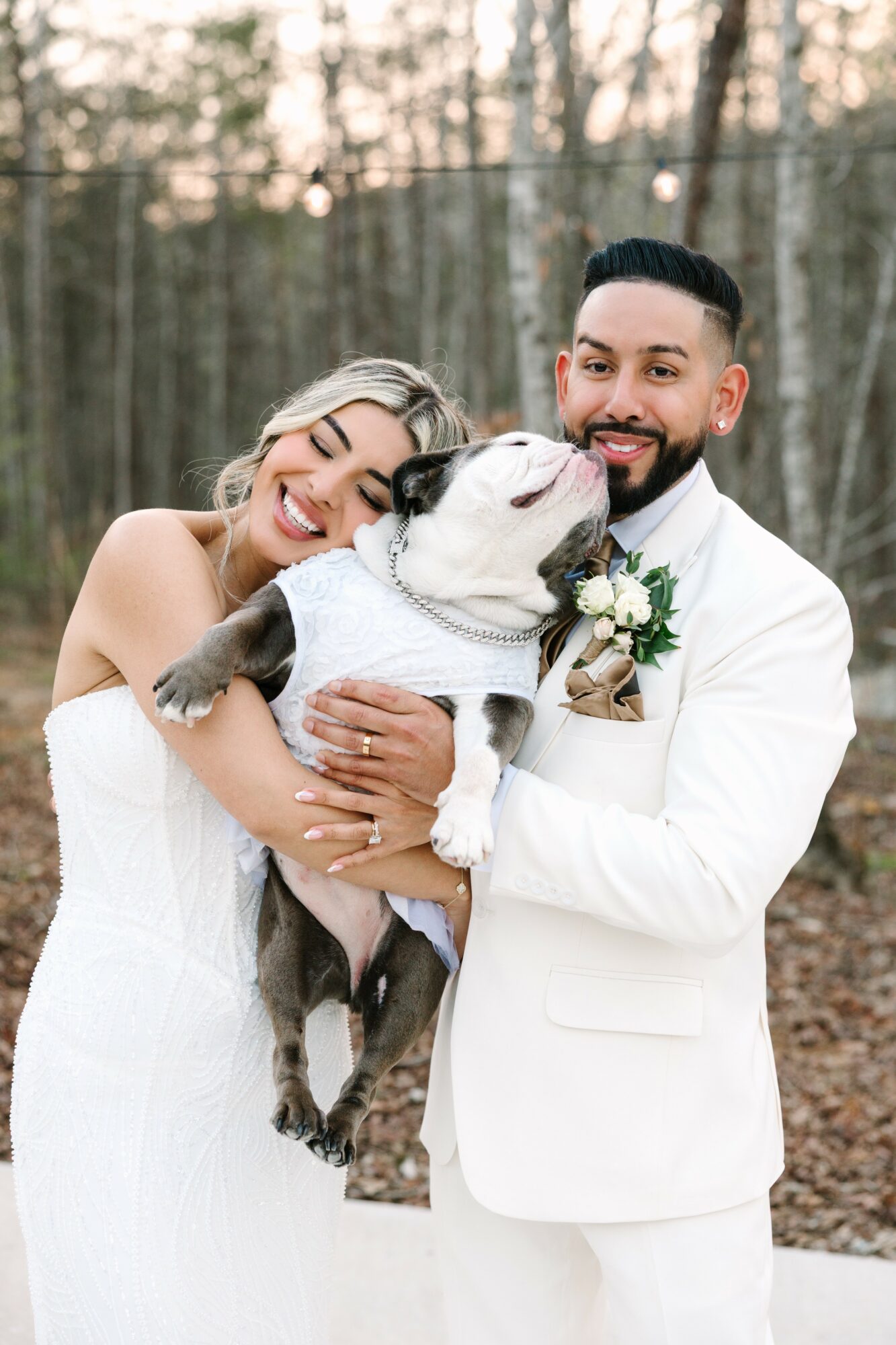 A couple in wedding attire holding a dog outdoors with trees in the background.