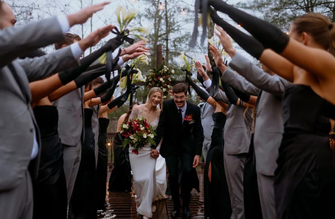 Bride and groom walking under archway of raised hands at wedding ceremony, surrounded by guests, outdoors, trees in background.