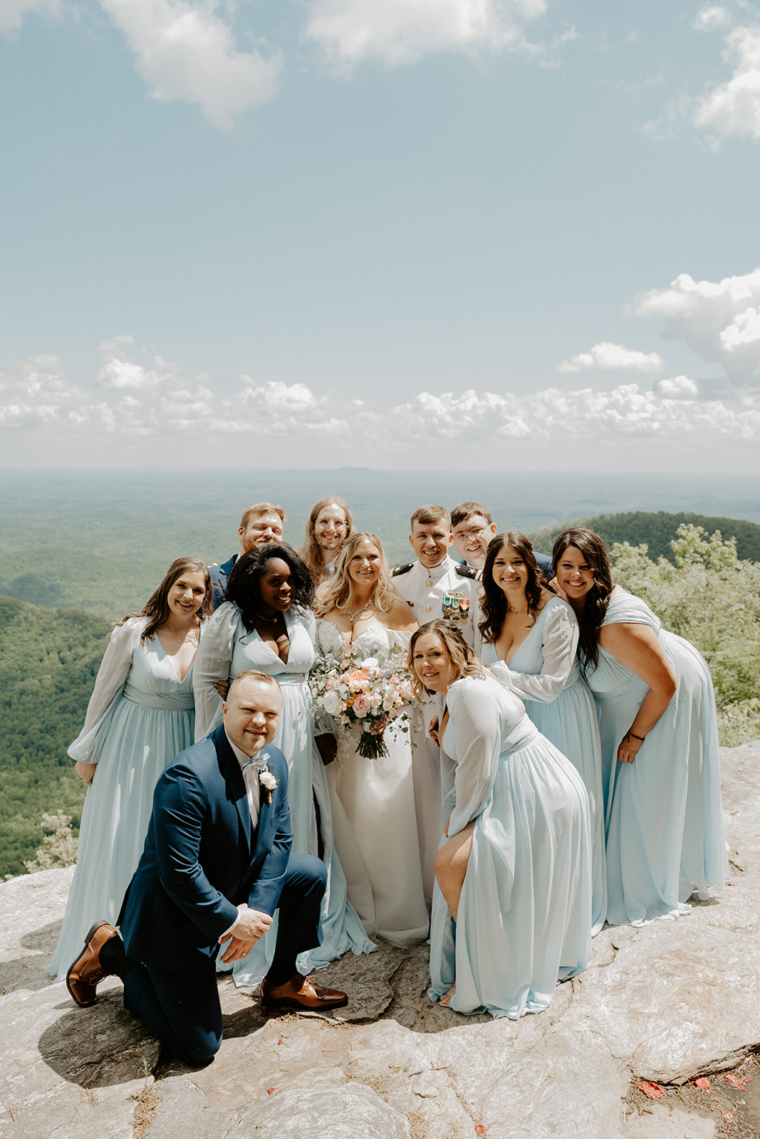 Group of people outdoors, some in formal attire, standing on a rocky surface with a landscape and sky in the background.