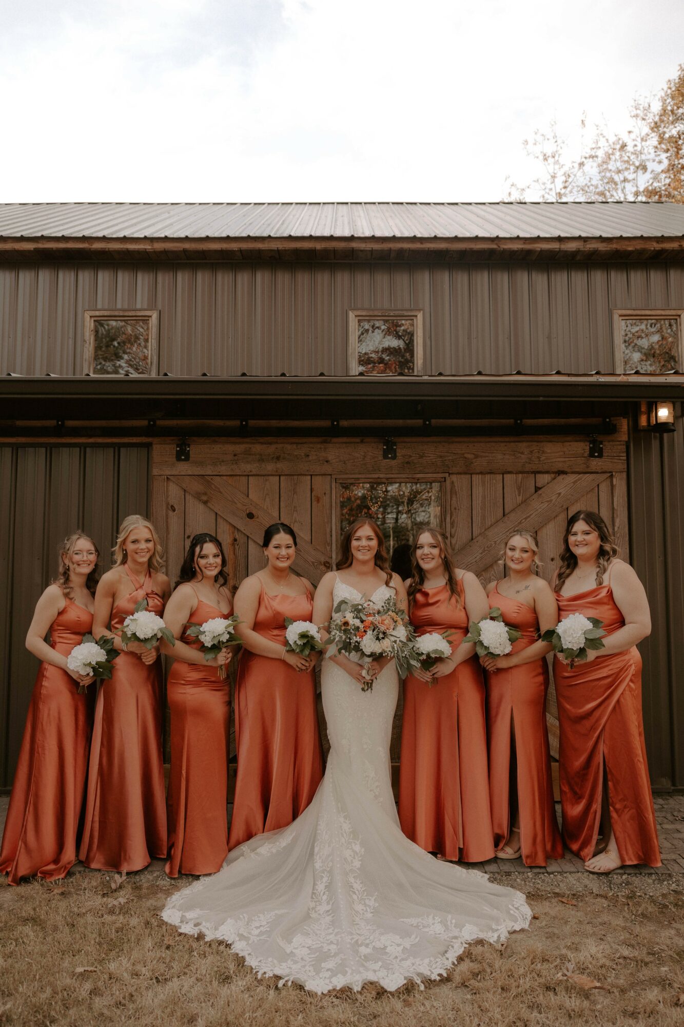 Group of seven women in formal dresses standing outdoors in front of a wooden building, holding bouquets.