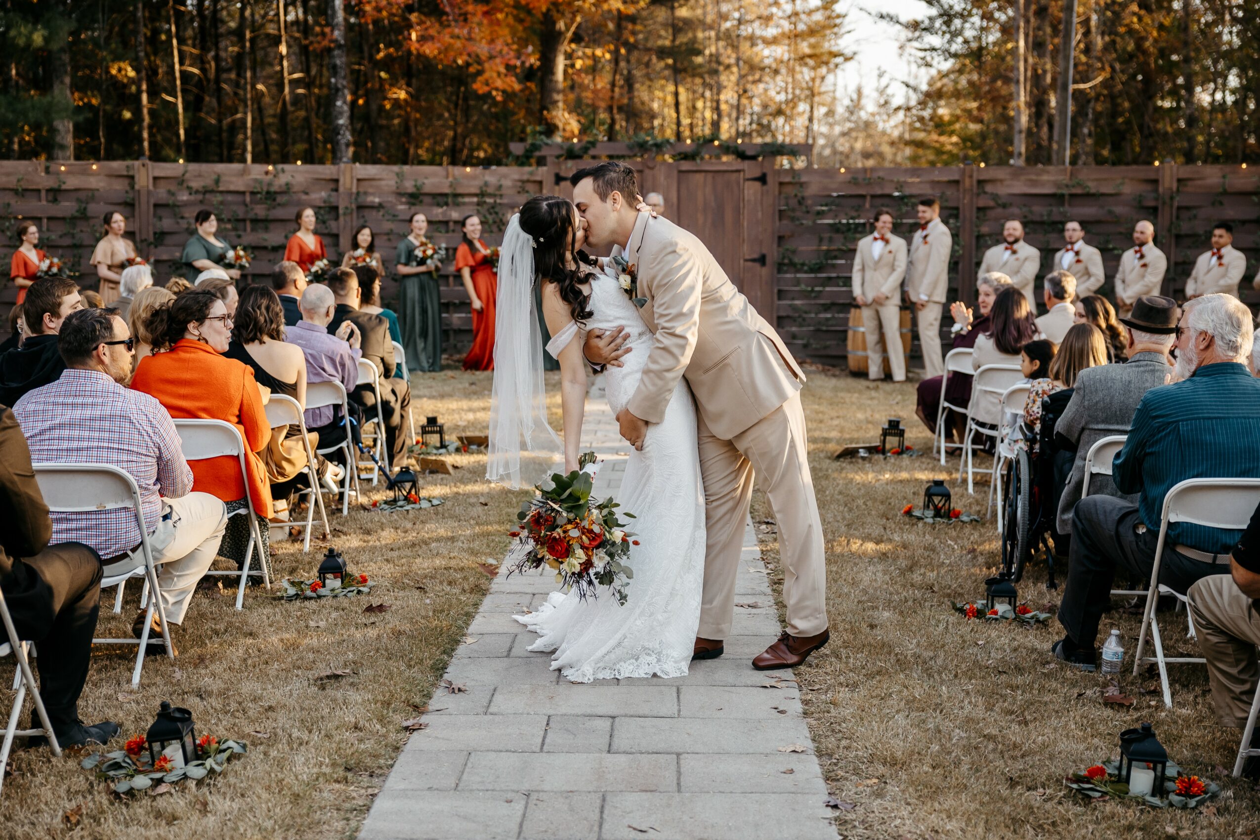 Bride and groom kiss on a wedding aisle outdoors, surrounded by seated guests and trees in the background.