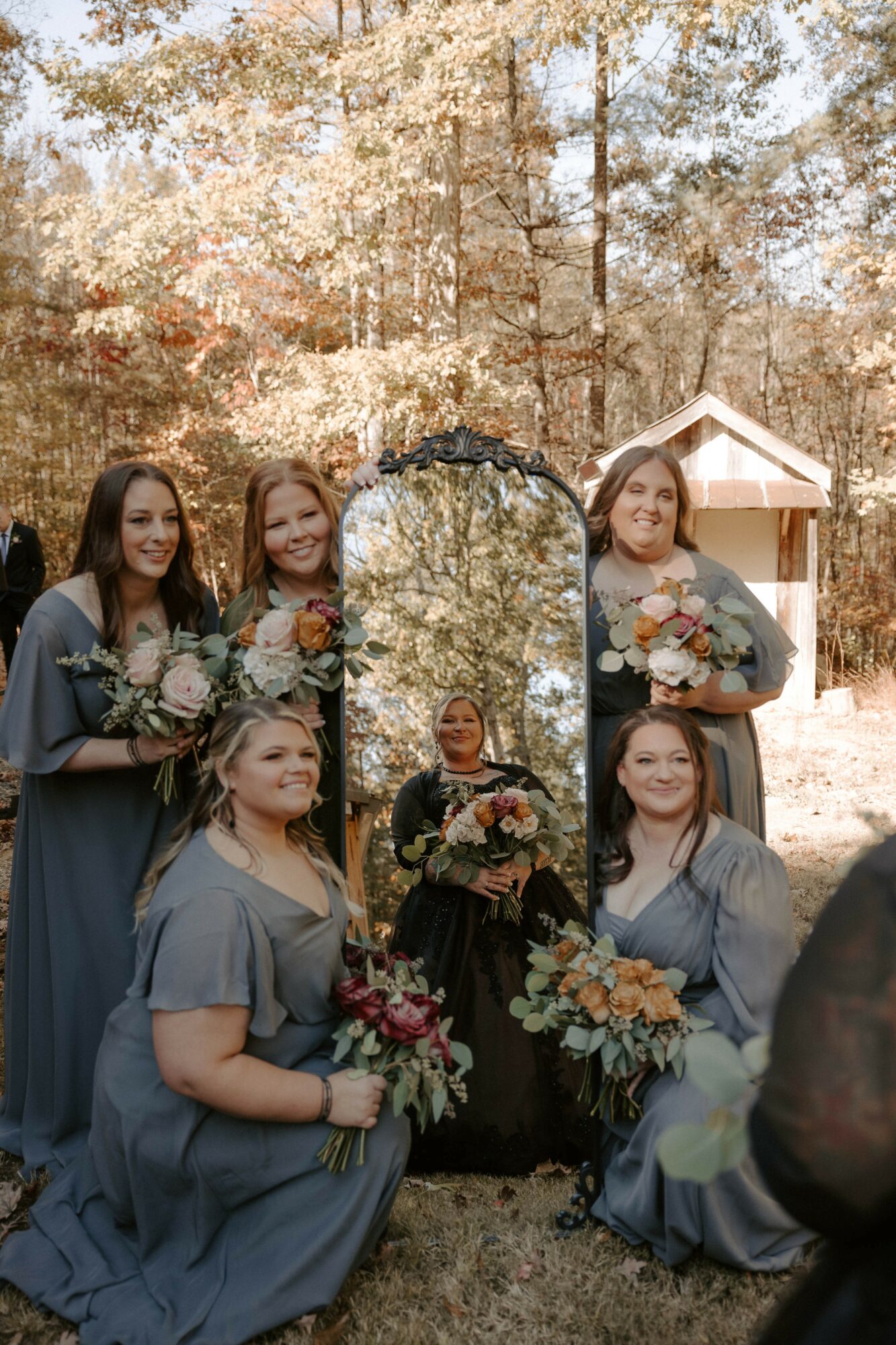 Five women in matching dresses holding bouquets outdoors, with trees and a small building in the background.