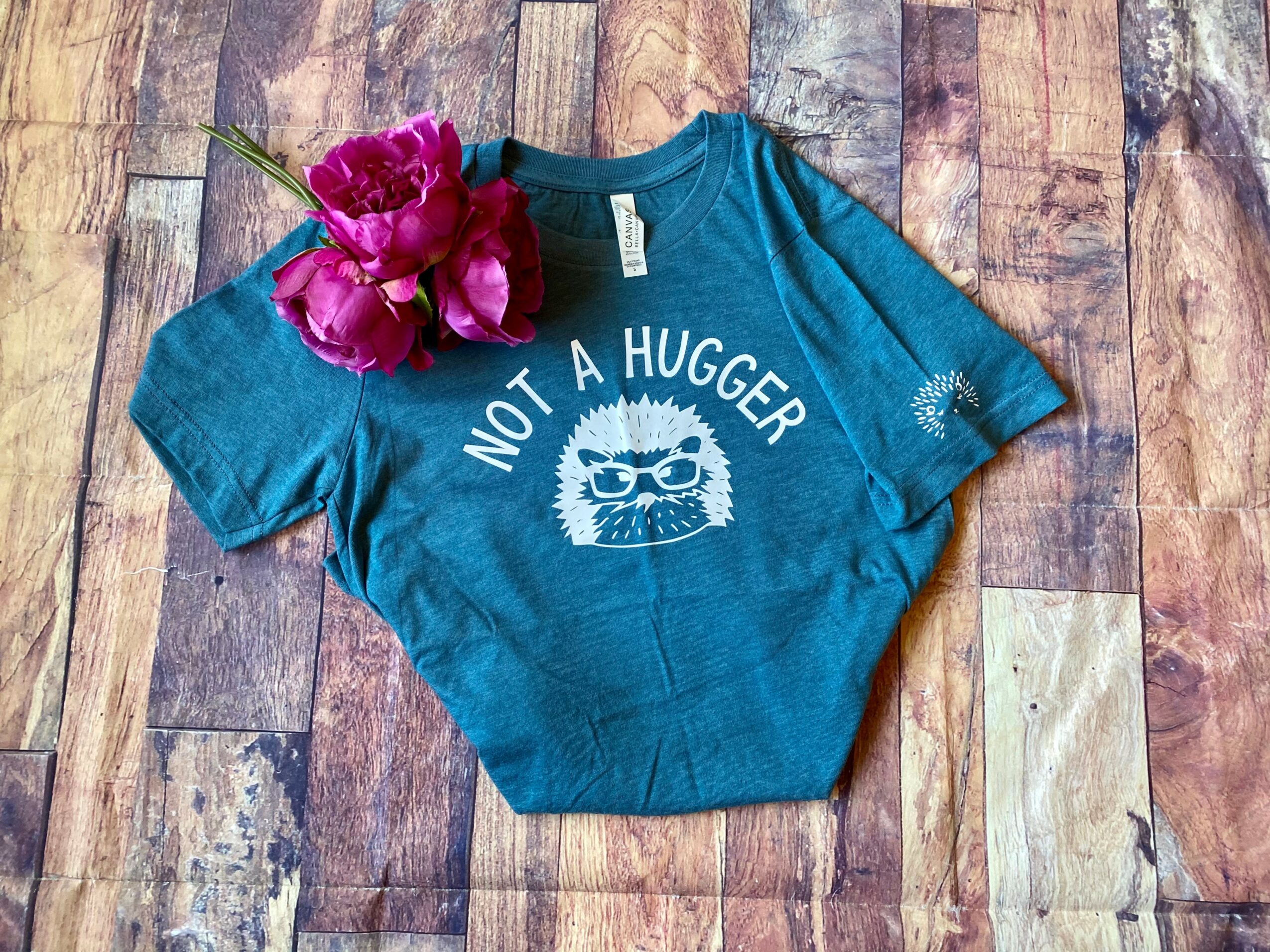 Blue children's T-shirt with a graphic and text, placed on a wooden surface with pink flowers on top.