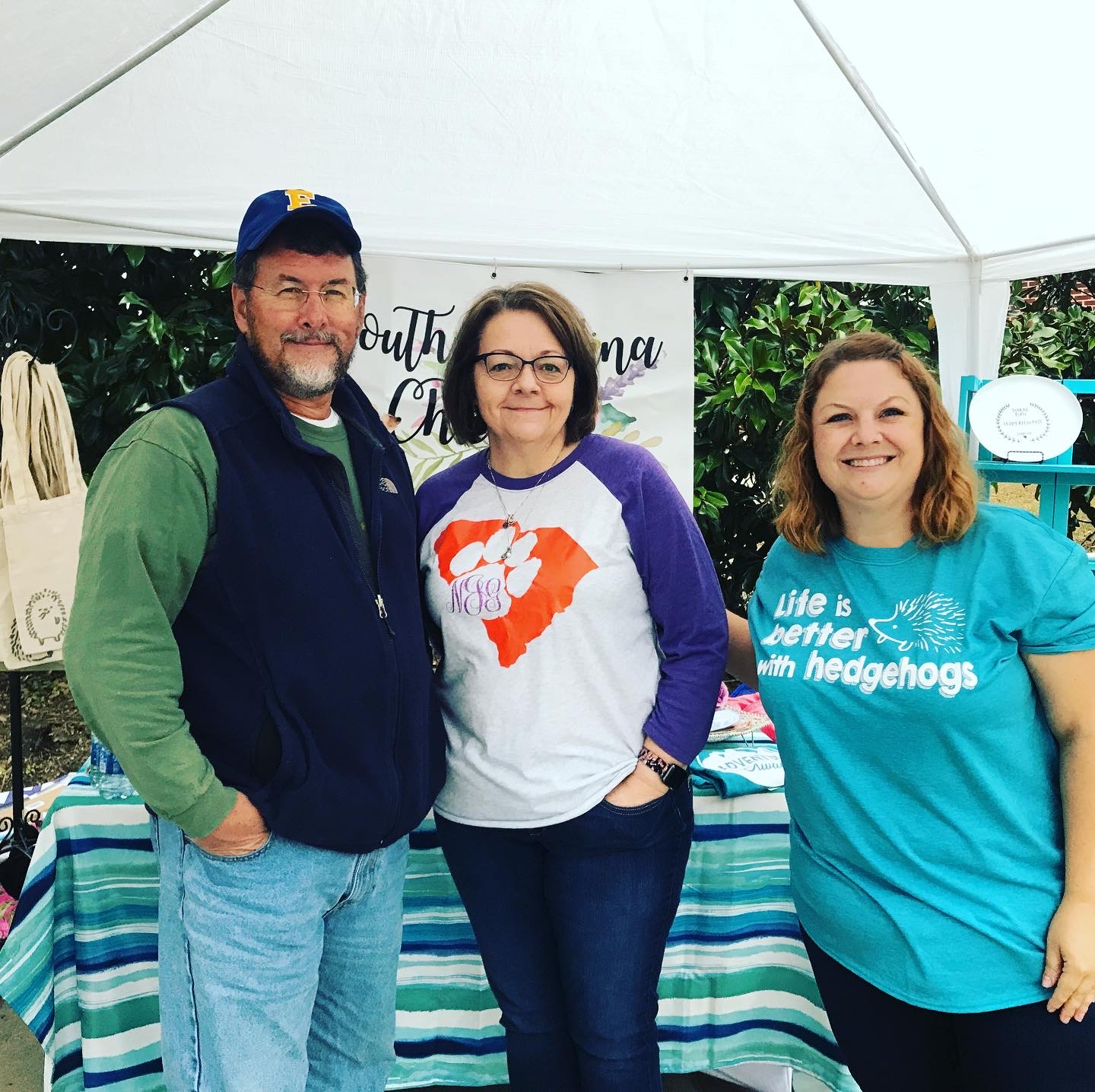 Three people standing outdoors under a white canopy, smiling, with a table and greenery in the background.