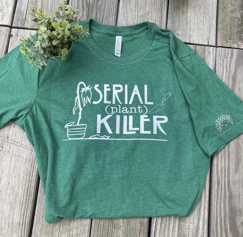 Green T-shirt with white text and plant graphic, laid on wooden surface, with a small plant on top.