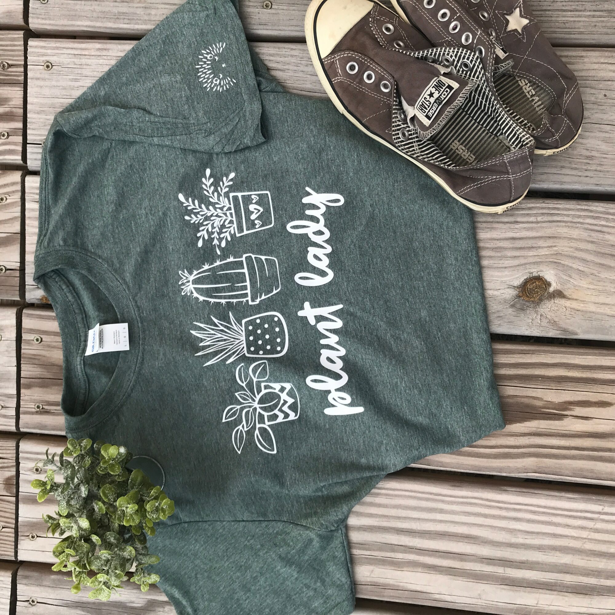 Gray T-shirt with white illustrations and text, placed on wooden surface with shoes and plant nearby.