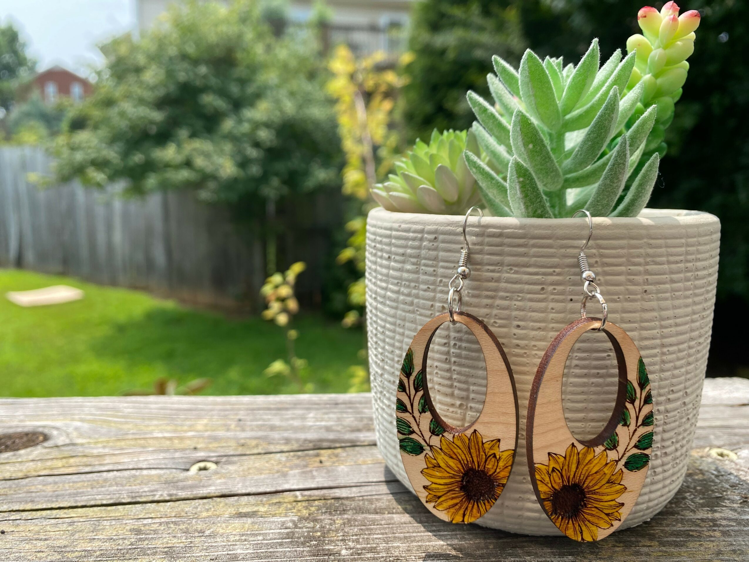 Earrings with sunflower design hanging in front of a potted succulent plant outdoors, wooden surface, garden background
