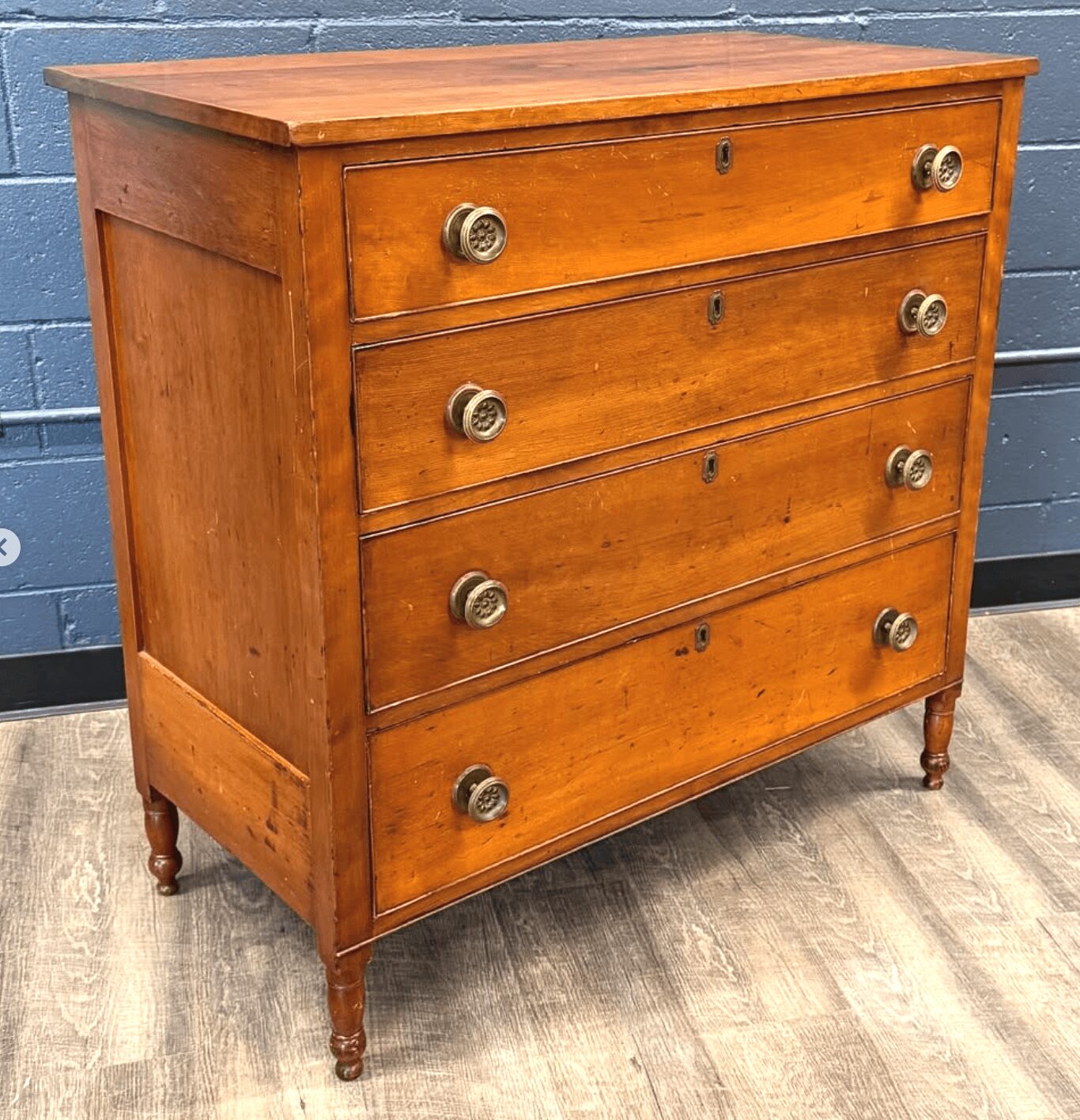 Wooden dresser with five drawers and round knobs, standing on four small turned legs, against a blue wall.