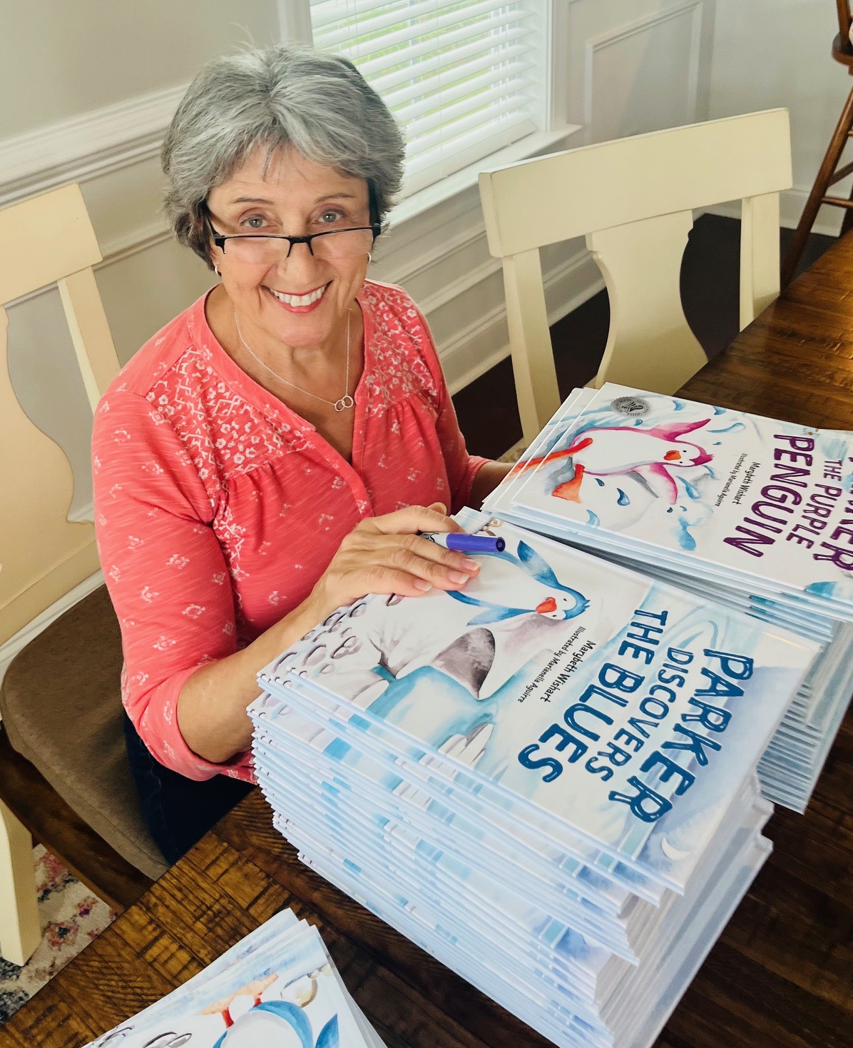 Smiling woman with glasses and gray hair sitting at a table with stacks of books about breastfeeding and the blues.