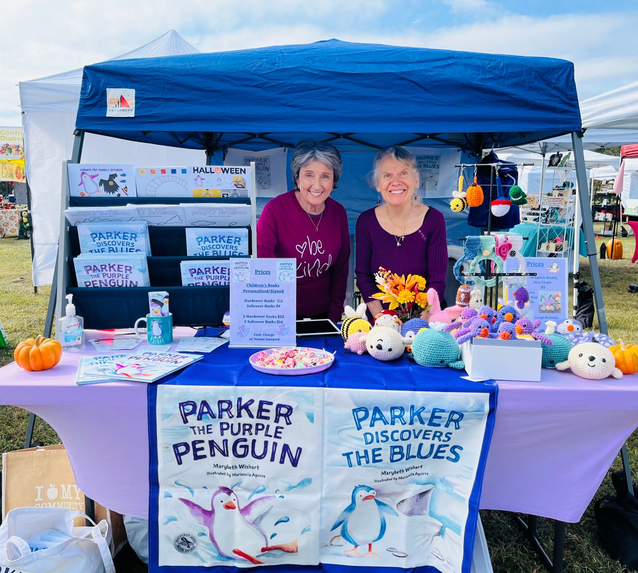Two women stand behind a booth with books, toys, and decorations at an outdoor event, smiling at the camera.