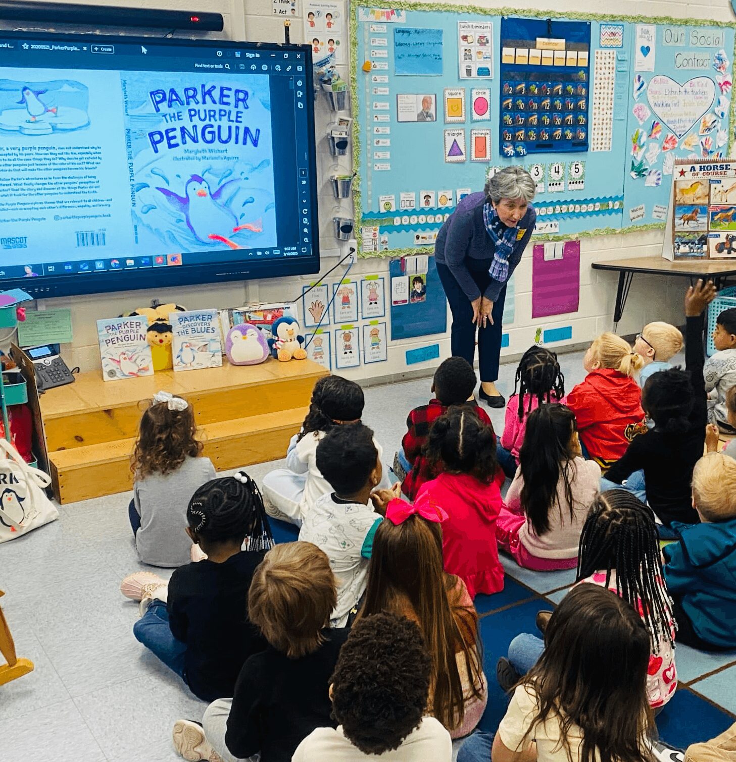 Teacher standing in front of students in a classroom, with a large screen displaying a book cover titled 'Parker the Purple Penguin'.