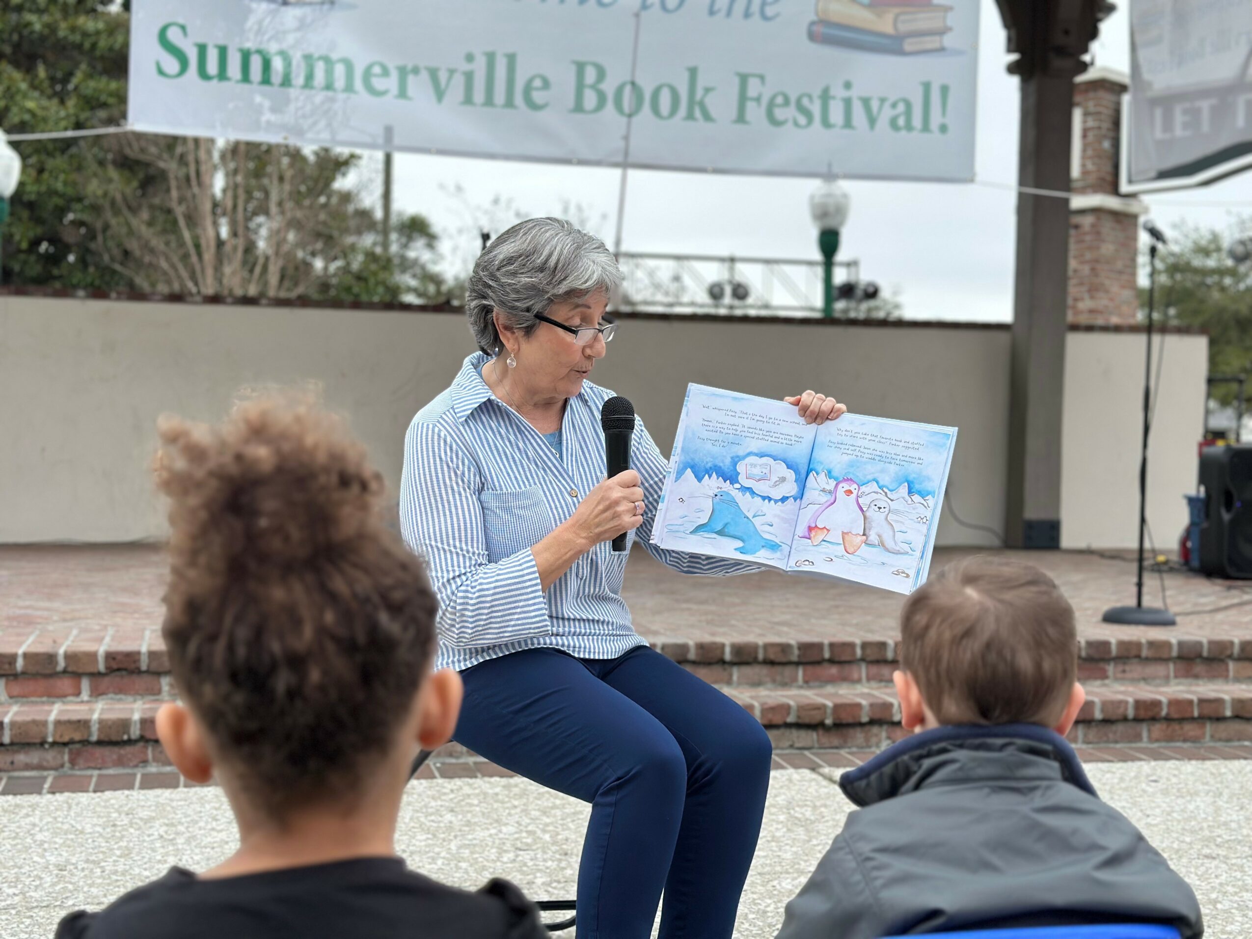 Woman reading a book to children outdoors at a book festival, holding a colorful illustrated book, with a banner in the background.
