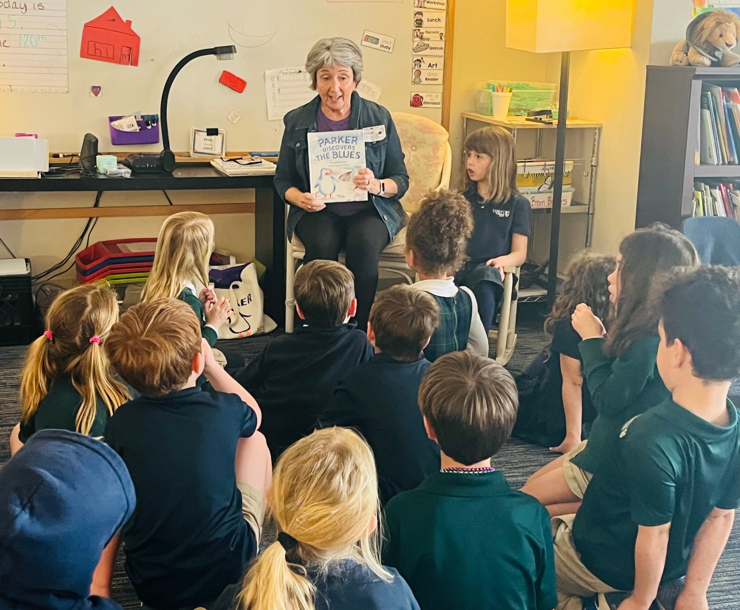 A woman reading a book to a group of children in a classroom, with bookshelves and educational materials visible.